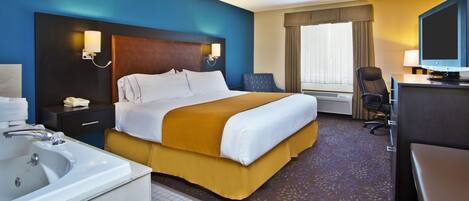A hotel room with a large bed, a desk with a computer, a TV, a bathroom with a sink and mirror, and a ceiling fan.