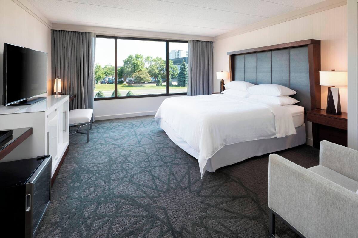 Habitación de hotel con cama, escritorio con televisor, una silla y ventana con cortinas.
