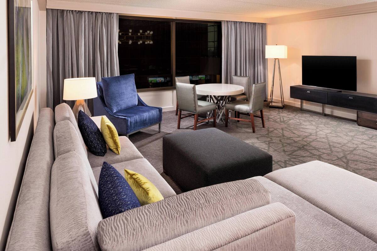 Habitación de hotel con un sofá grande, una mesa de comedor con sillas, un televisor de pantalla plana y una lámpara.