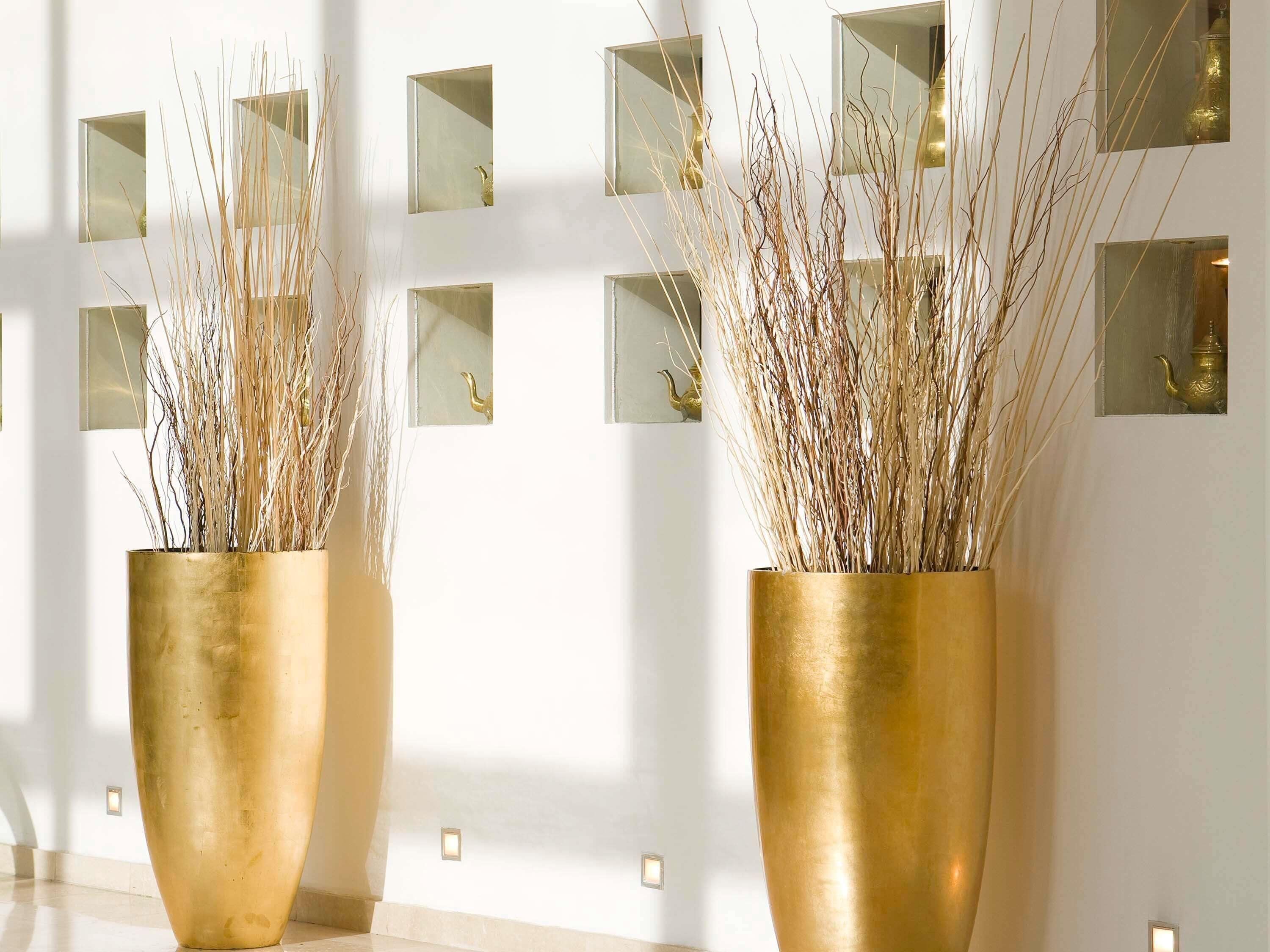 Two large gold vases with dried plants on a polished floor, set against a wall with multiple square mirrors and small lights.