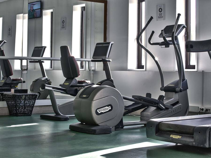 Un gimnasio con diferentes máquinas de ejercicio, una pantalla de televisión y una ventana con persianas.