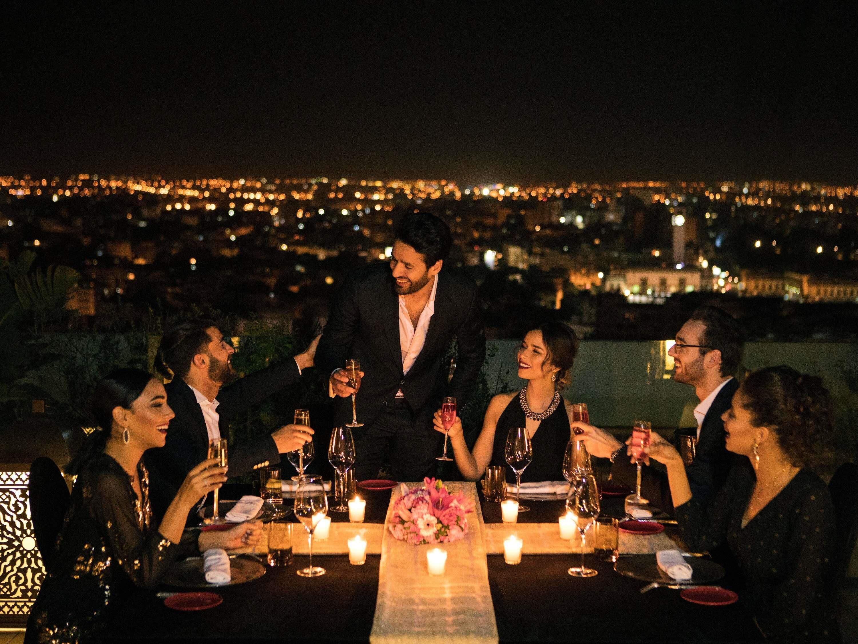 Un grupo de personas cenando en una mesa al aire libre con velas, con vistas a un paisaje urbano nocturno.