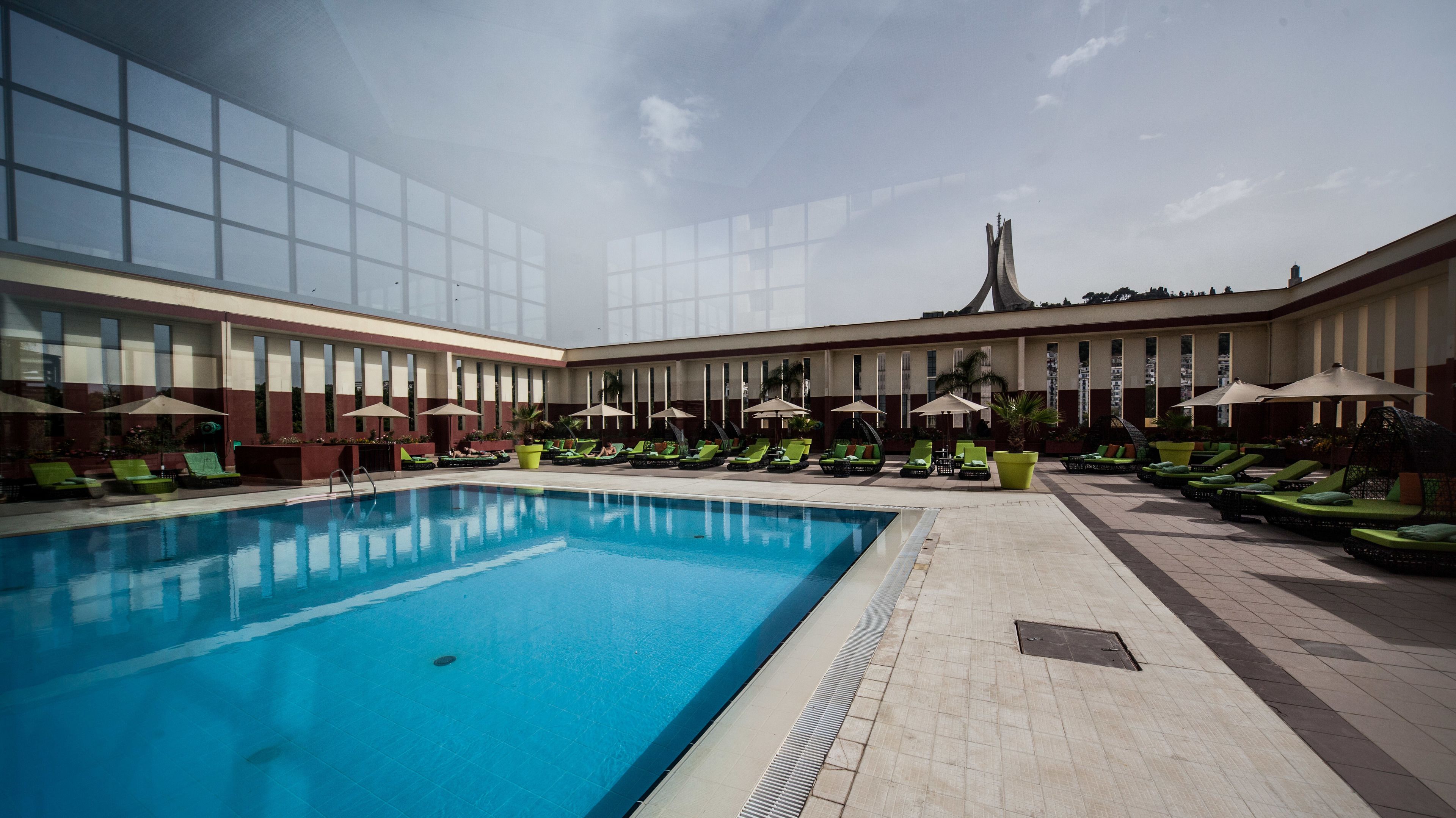 Zona de piscina con sillones de descanso y sombrillas, cielo despejado y un edificio con grandes ventanales al fondo.