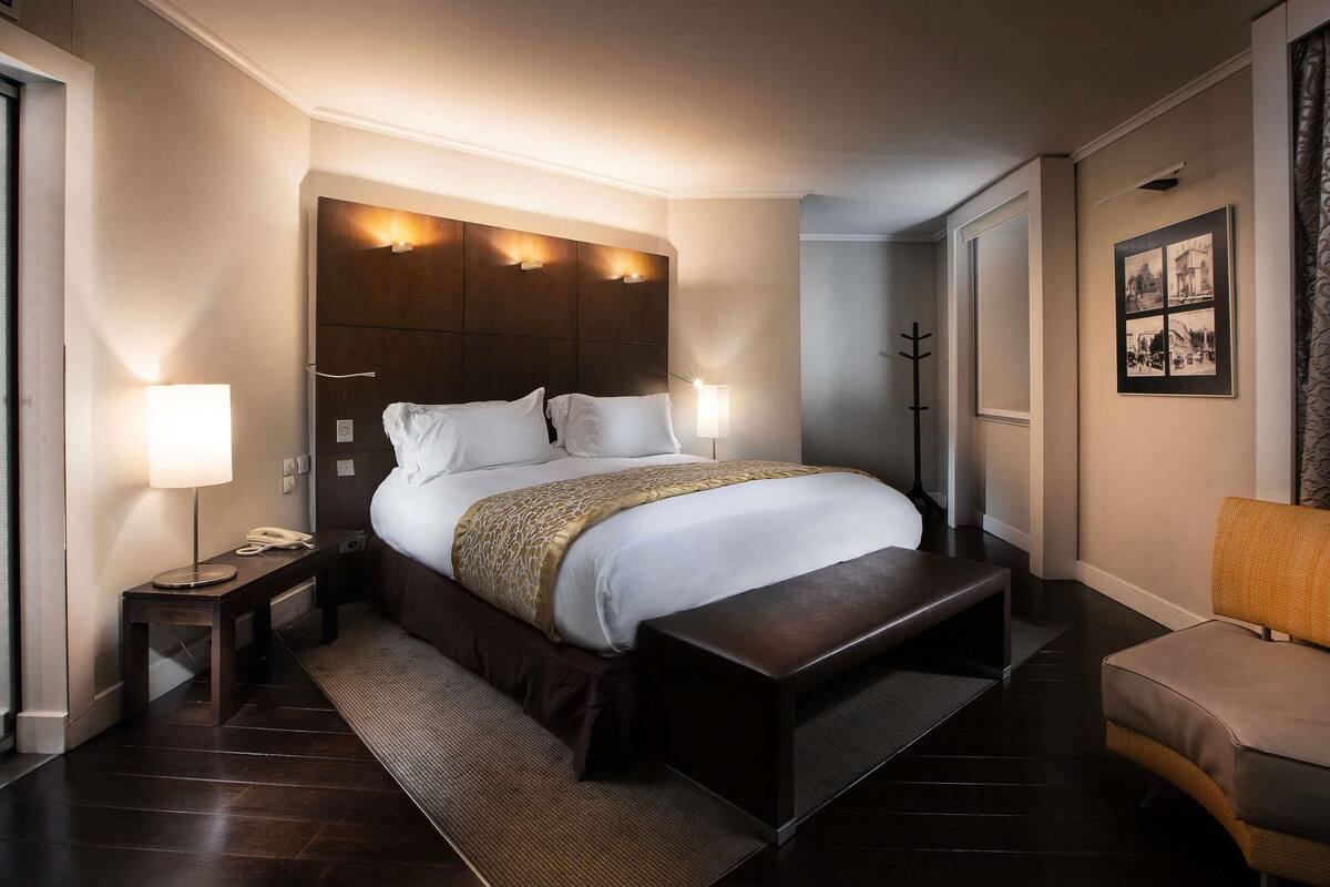 A hotel room with a large bed, bedside lamps, a bench, and a chair.