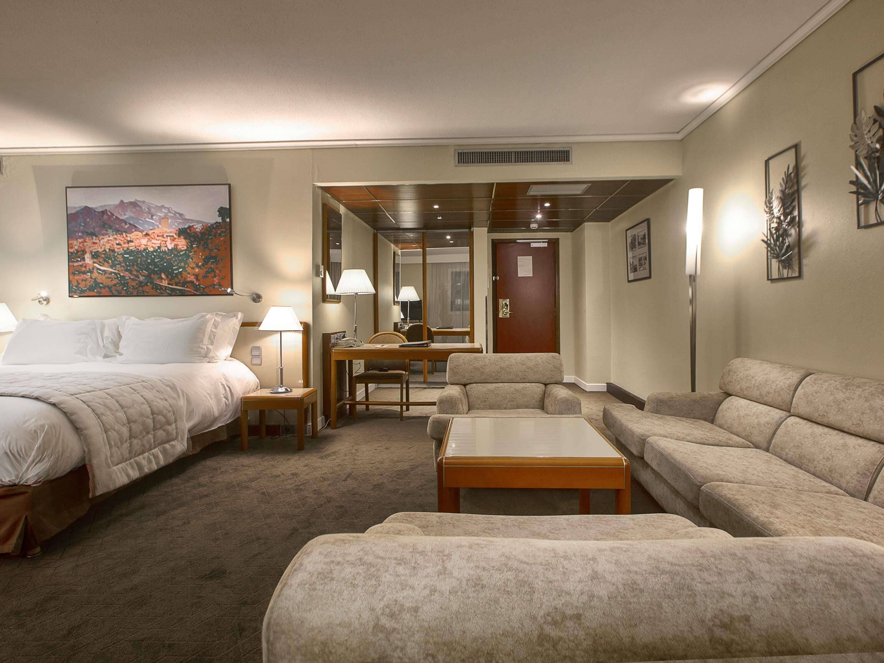 A hotel room with a bed, two armchairs, a sofa, a desk, and a painting of a mountain landscape.
