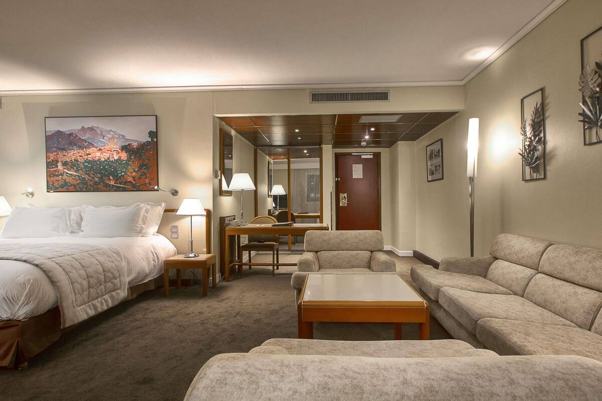 A hotel room with a bed, two armchairs, a sofa, a desk, and a painting of a mountain landscape.
