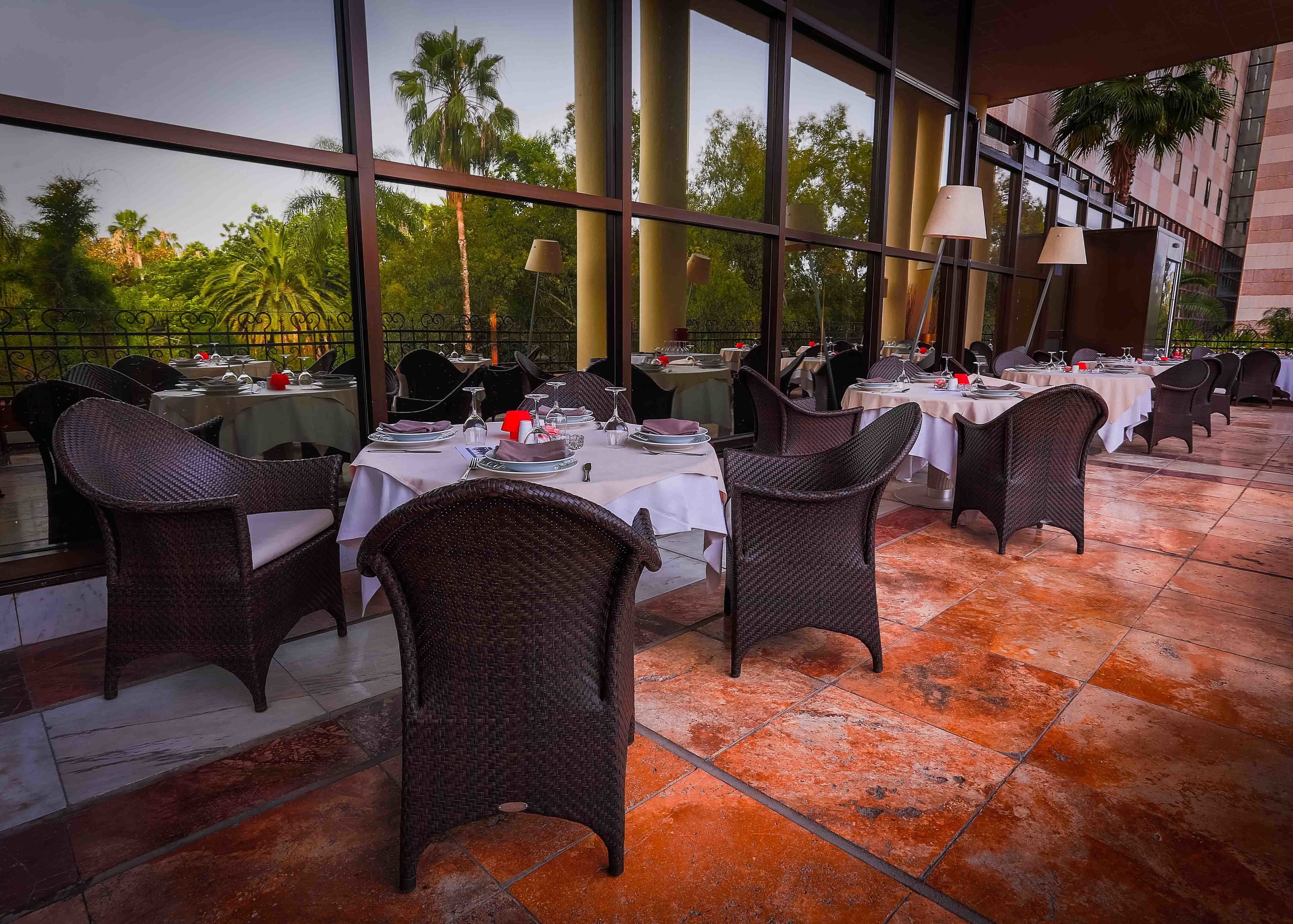 An outdoor dining area with wicker chairs and tables, large windows, and a view of palm trees.