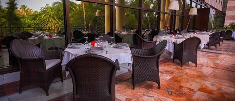 An outdoor dining area with wicker chairs and tables, large windows, and a view of palm trees.