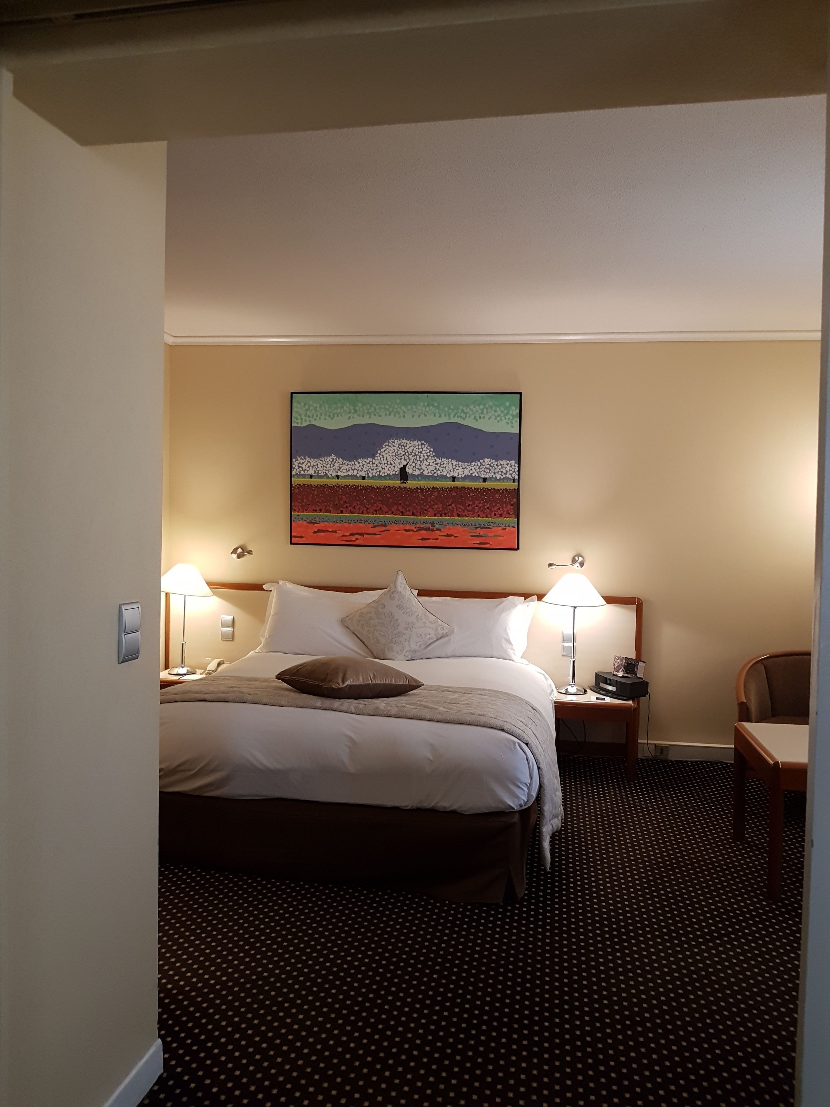Ein Hotelzimmer mit einem Bett, zwei Nachttischlampen, einem Nachttisch, einem Schreibtisch mit Stuhl und einem Bild an der Wand.