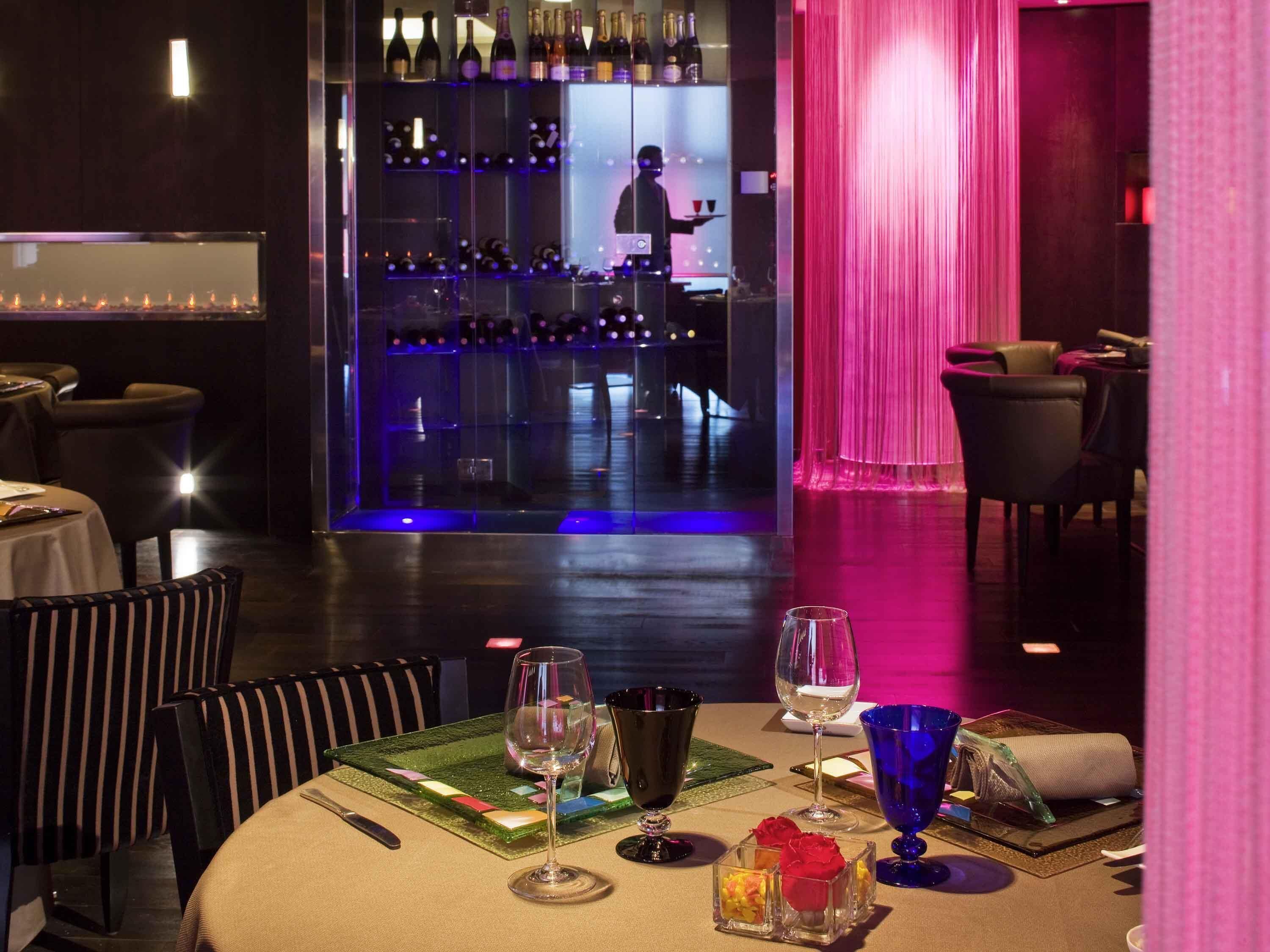 Un restaurante con zona de bar, mesas con copas de vino y un camarero.
