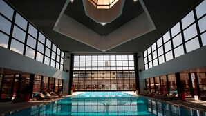 An indoor swimming pool with clear glass walls, sunlit interior, and lounge chairs along the sides. - Sofitel Algiers Hamma Garden (Algiers)