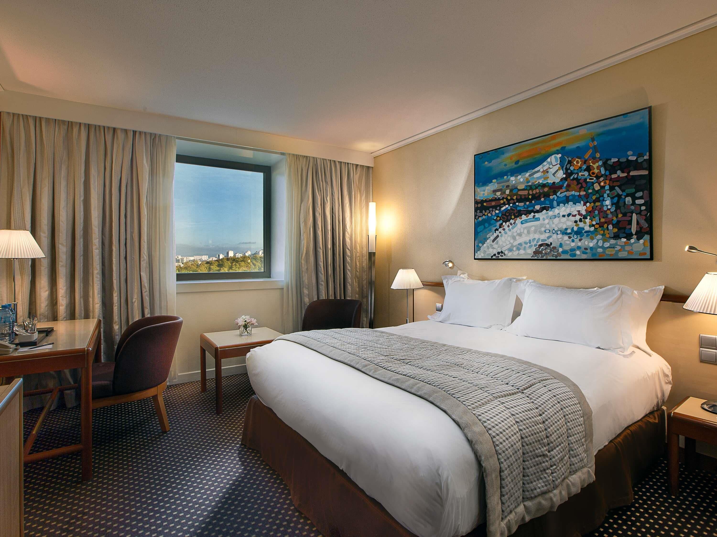 A hotel room with a bed, a desk, a chair, a window with curtains, a painting on the wall, and a patterned carpet.