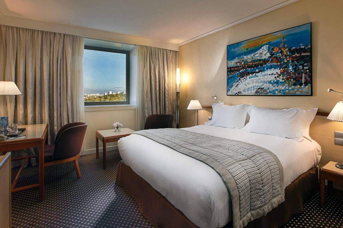 A hotel room with a bed, a desk, a chair, a window with curtains, a painting on the wall, and a patterned carpet.