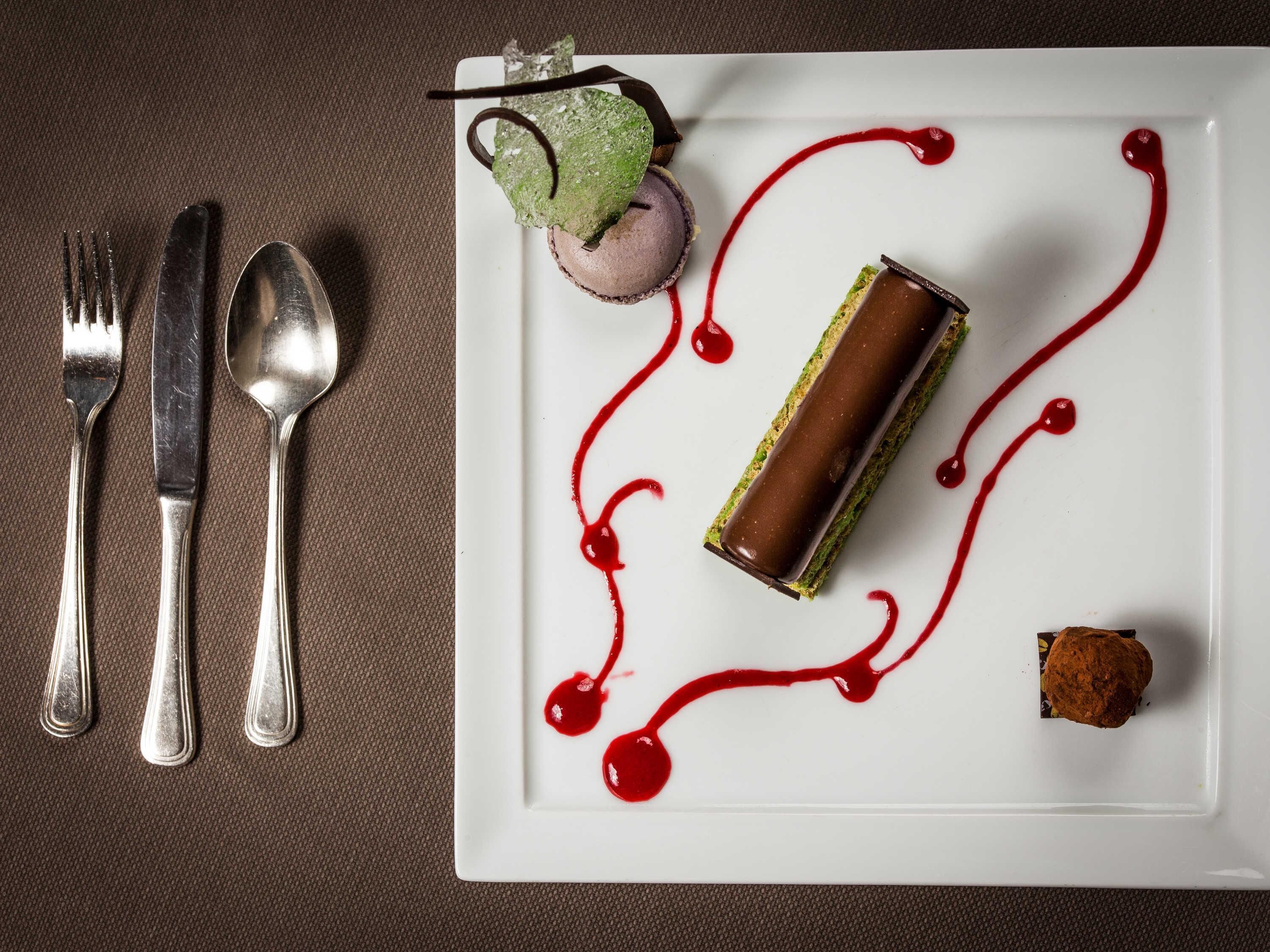 A dessert plate with a chocolate roll, a green and white dessert, and a small brown item, accompanied by a fork, knife, and spoon on a textured surface.