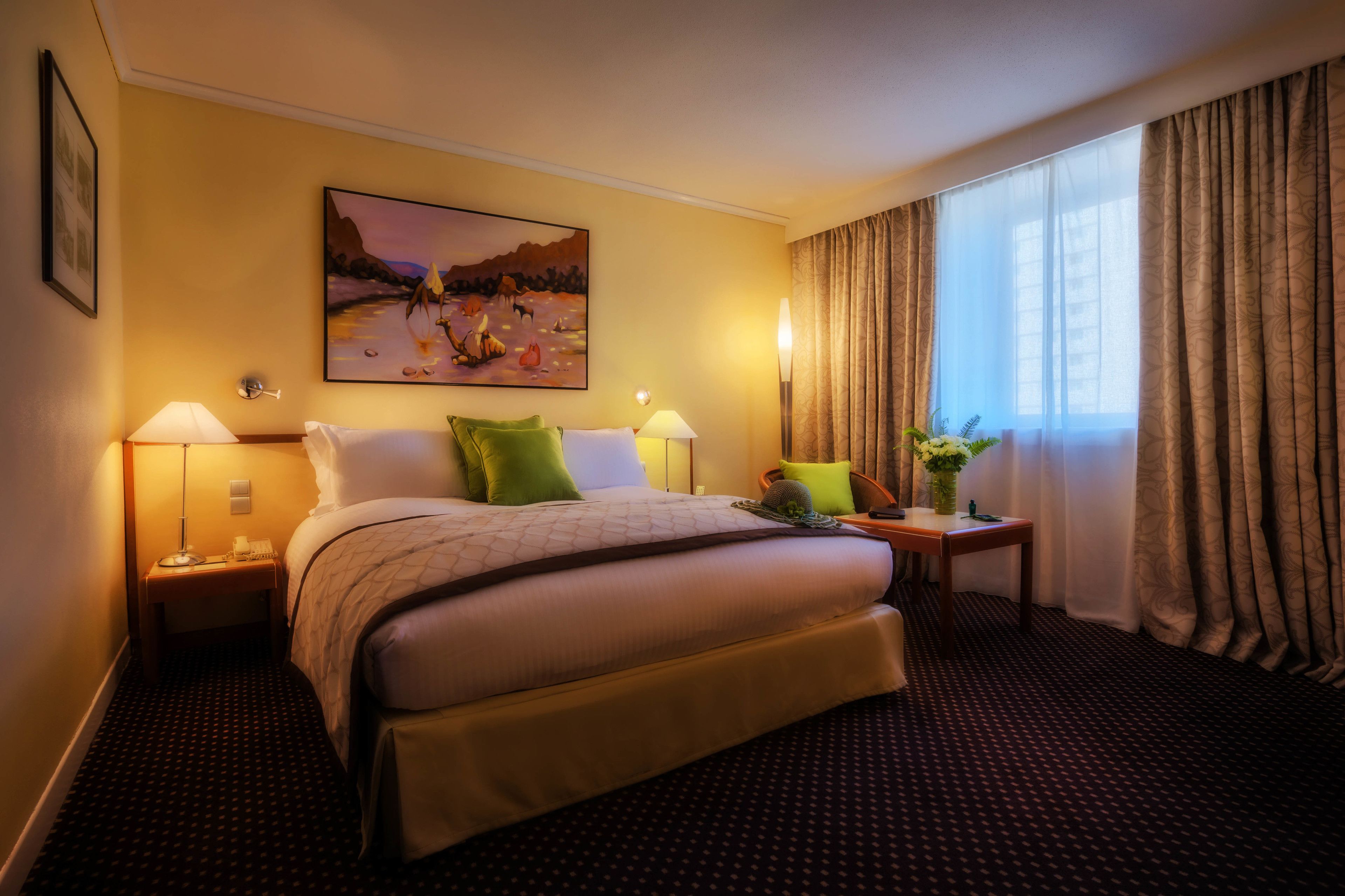 A hotel room with a bed, two bedside tables, a nightstand, a painting, a window with curtains, and a patterned carpet.