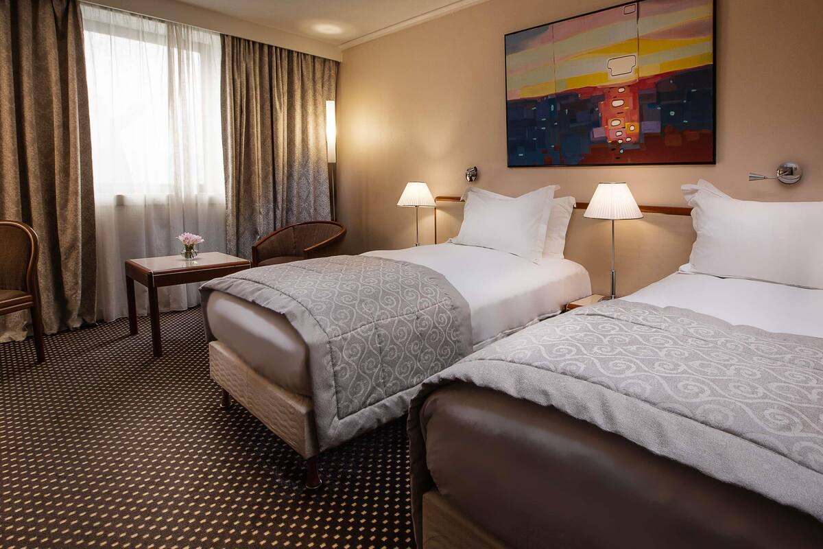A hotel room with two beds, a small table with a chair, a lamp, and a painting on the wall.