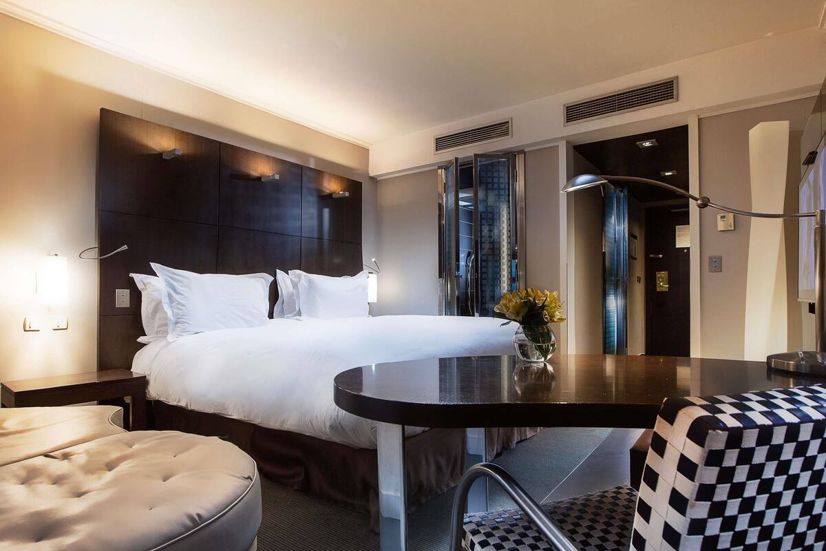 A hotel room with a large bed, a round table, a checkered chair, and a small vase with flowers.