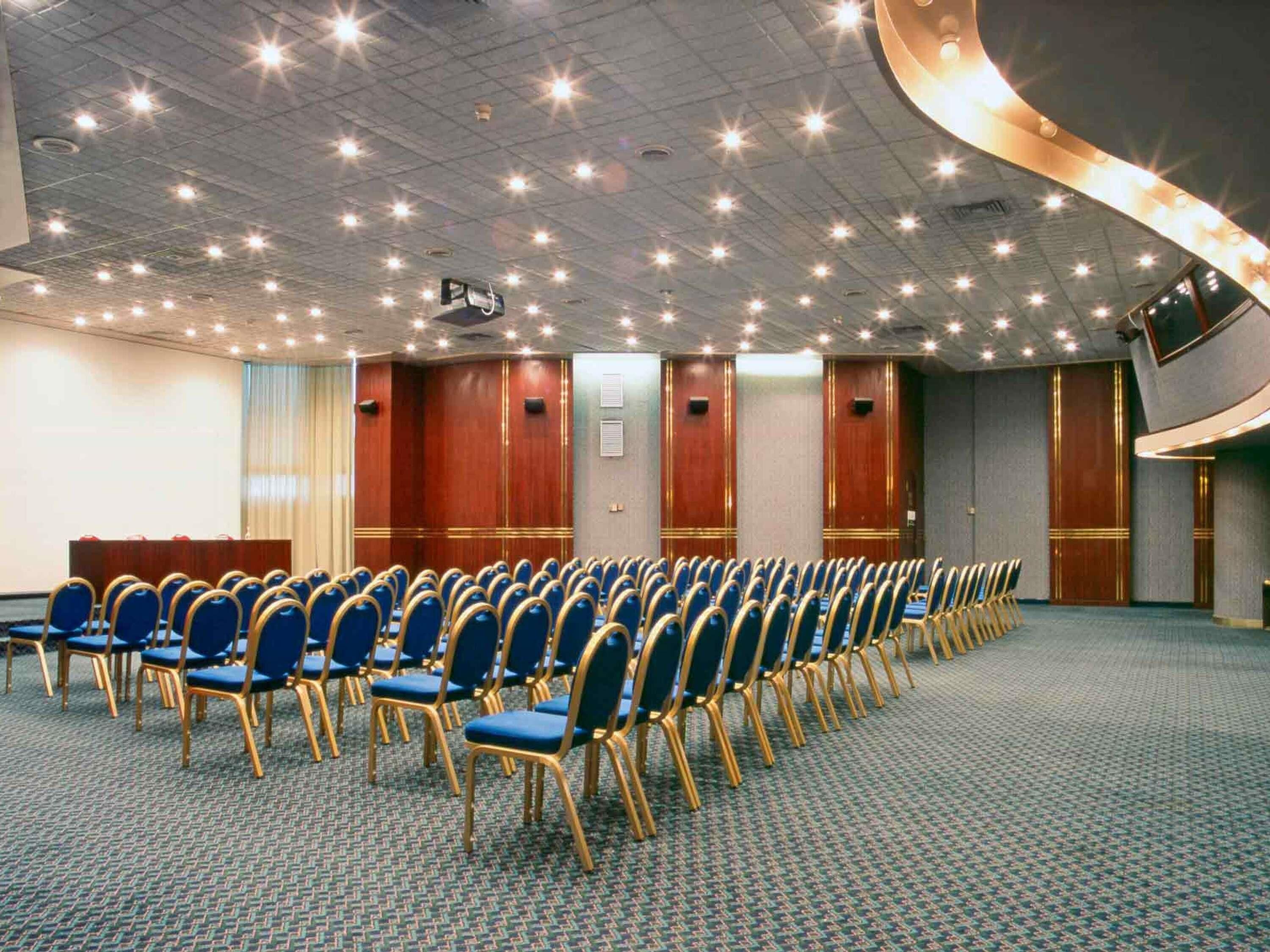 Un salón de conferencias con hileras de sillas azules, un atril y un techo curvo con iluminación empotrada.