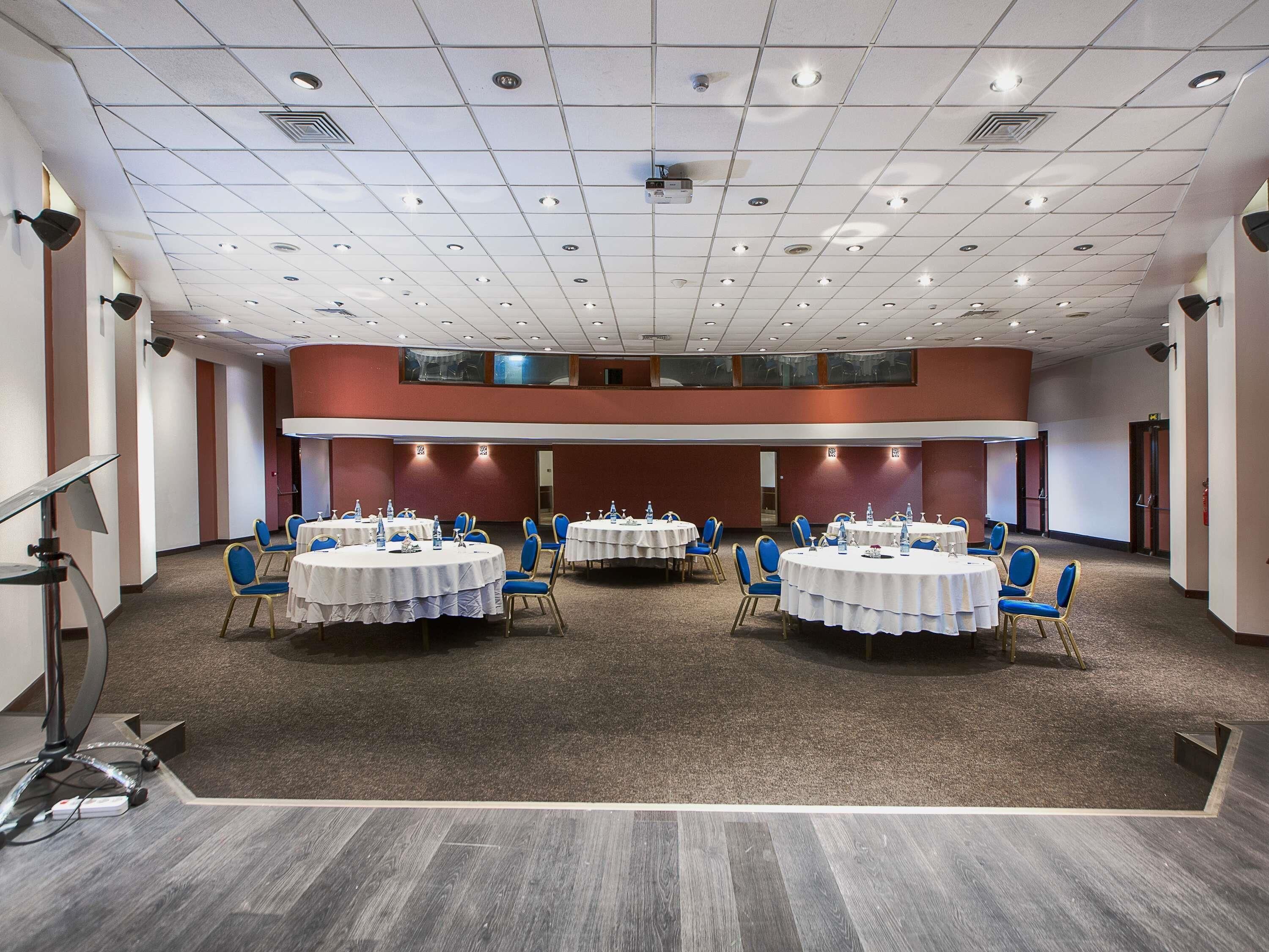 Une salle de conférence dotée de plusieurs tables et chaises, d’un sol recouvert de moquette et d’un plafond haut avec des luminaires encastrés.