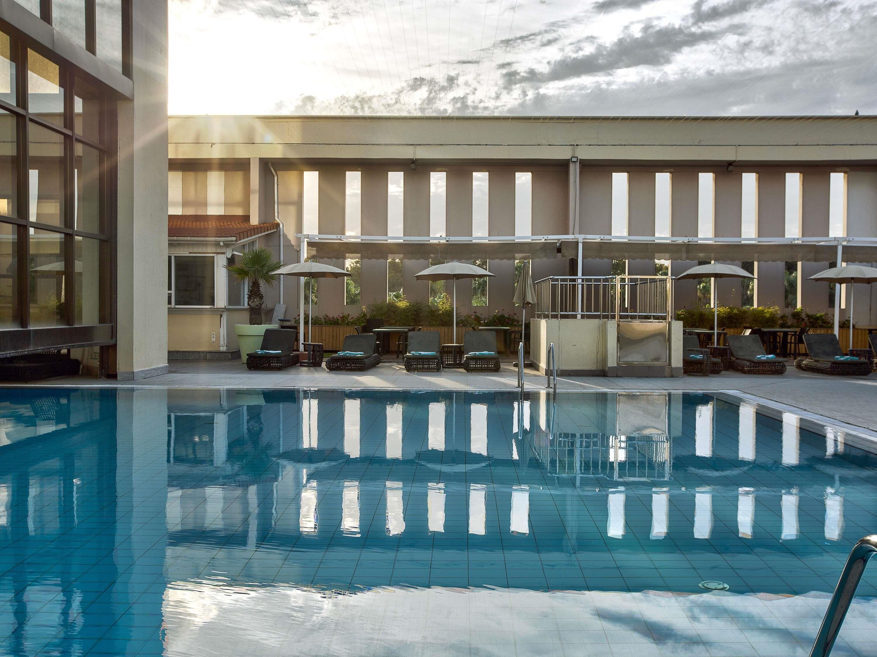 Una piscina con sillones de descanso y sombrillas, rodeada por un edificio con ventanas amplias y suelo de baldosas.