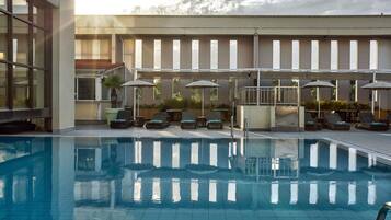 A swimming pool with lounge chairs and umbrellas, surrounded by a building with large windows and a tiled floor.