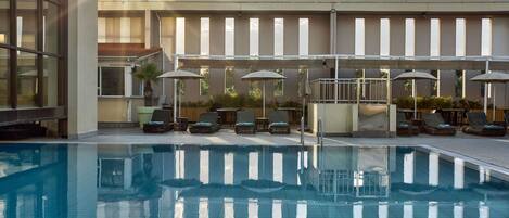 Una piscina con sillones de descanso y sombrillas, rodeada por un edificio con ventanas amplias y suelo de baldosas.