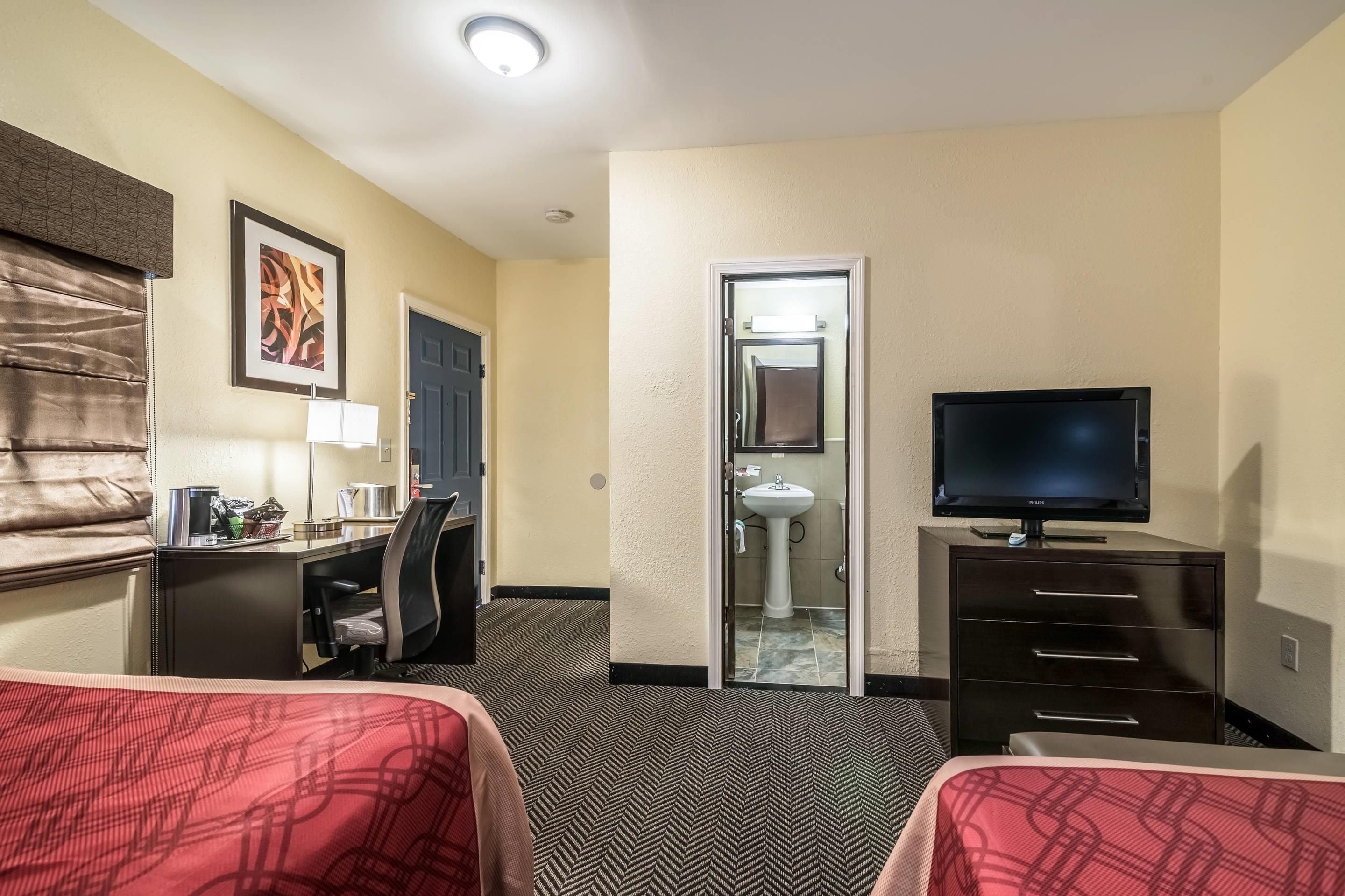 Quarto de hotel com duas camas, uma escrivaninha, uma cadeira, televisão e banheiro.