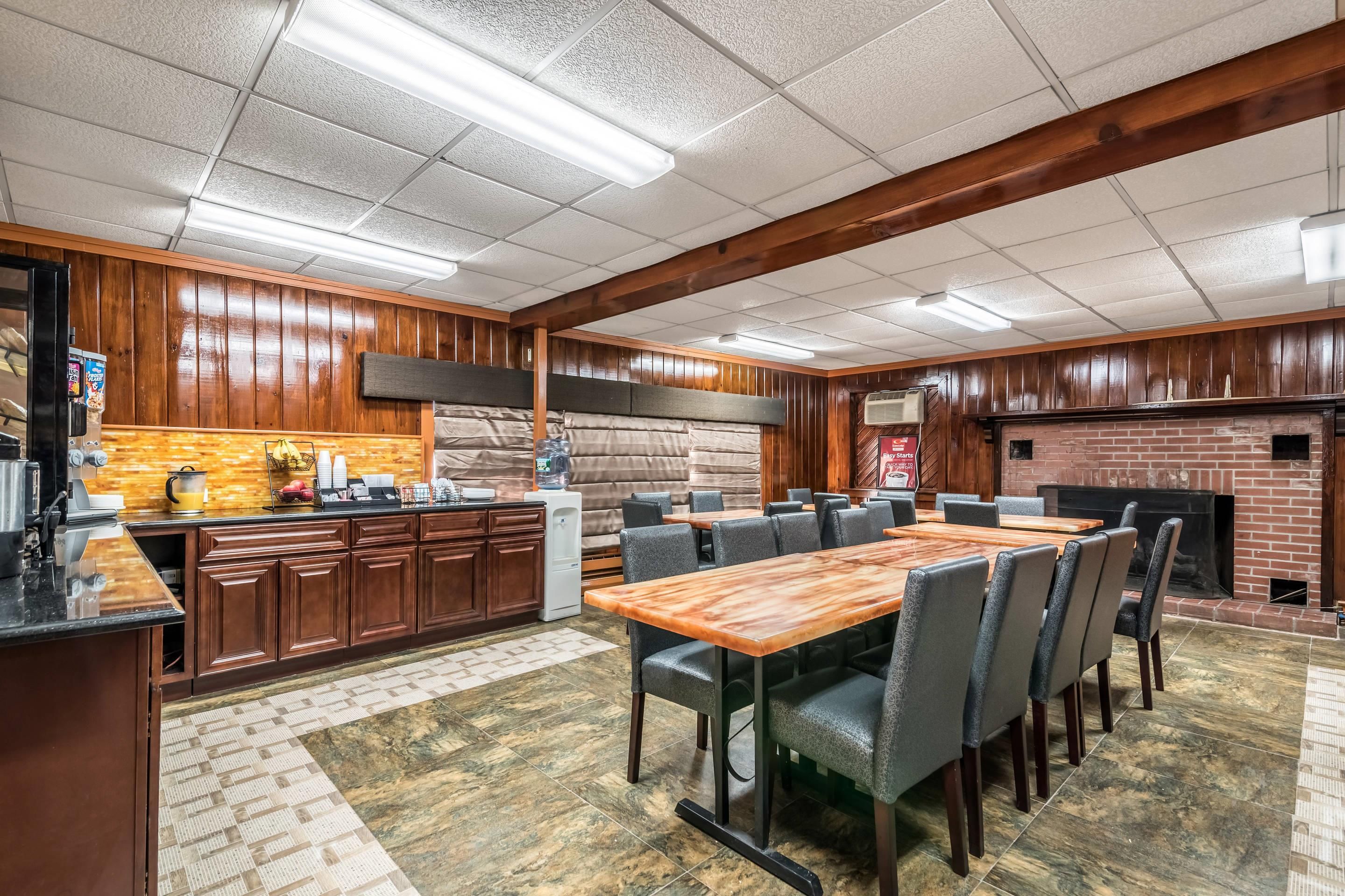 A restaurant with a long wooden table, cushioned chairs, a kitchen area, and a brick fireplace.