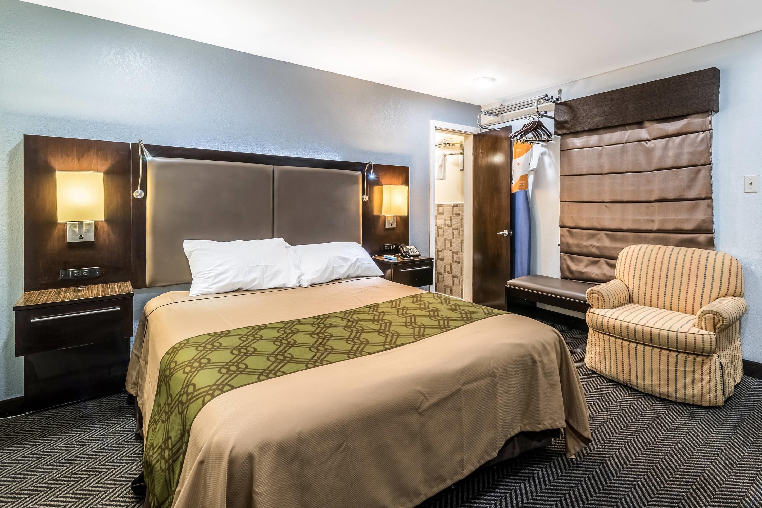 Habitación de hotel con cama, mesitas de noche, una silla, armario y baño visible a través de una puerta abierta.