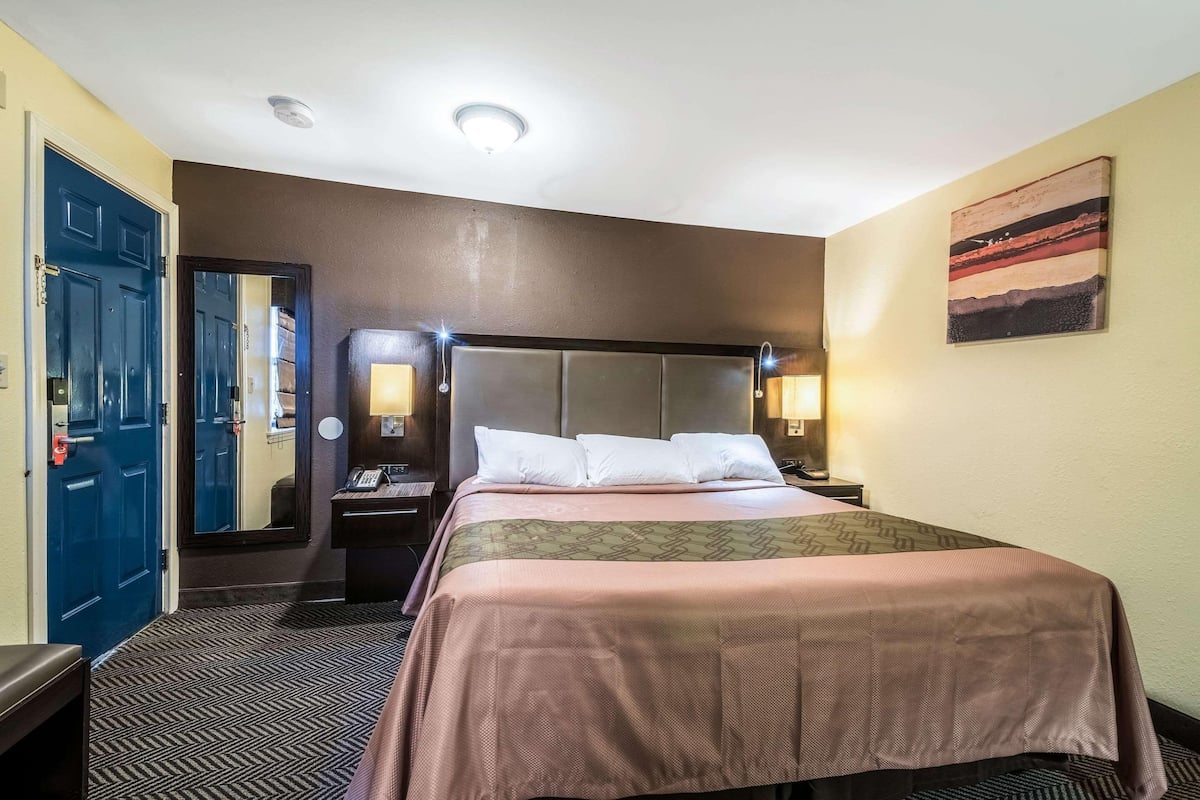 Una habitación de hotel con cama, mesita de noche, teléfono, espejo y un cuadro en la pared.