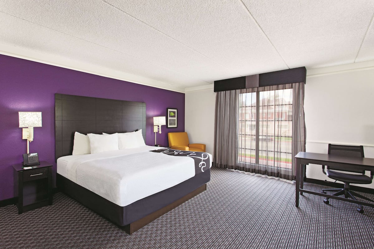 A hotel room with a large bed, a desk, and a chair. There is a window with curtains and a view of the outside.