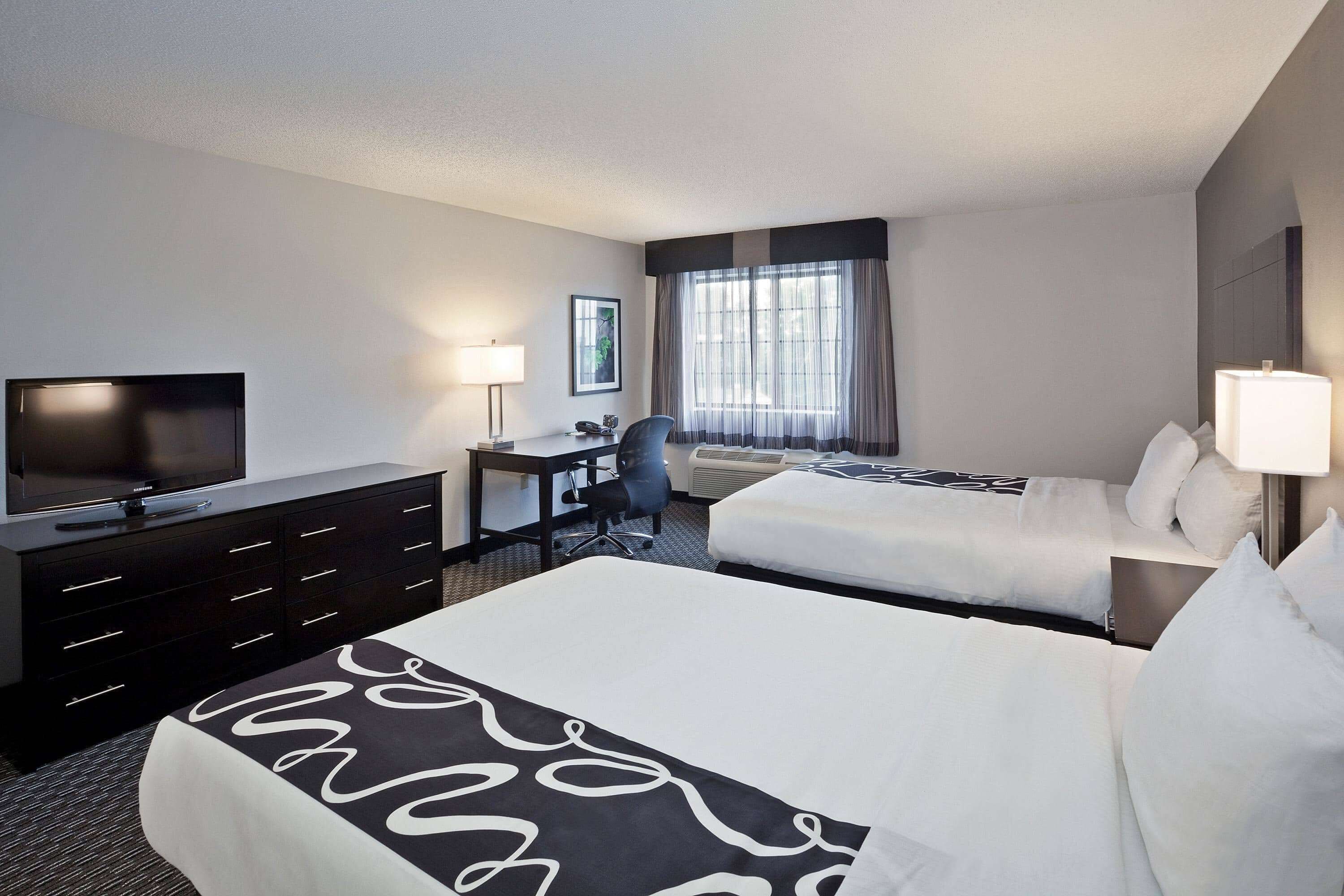 Une chambre d’hôtel comprenant un lit, un bureau avec une chaise, une télévision et une fenêtre avec des rideaux.