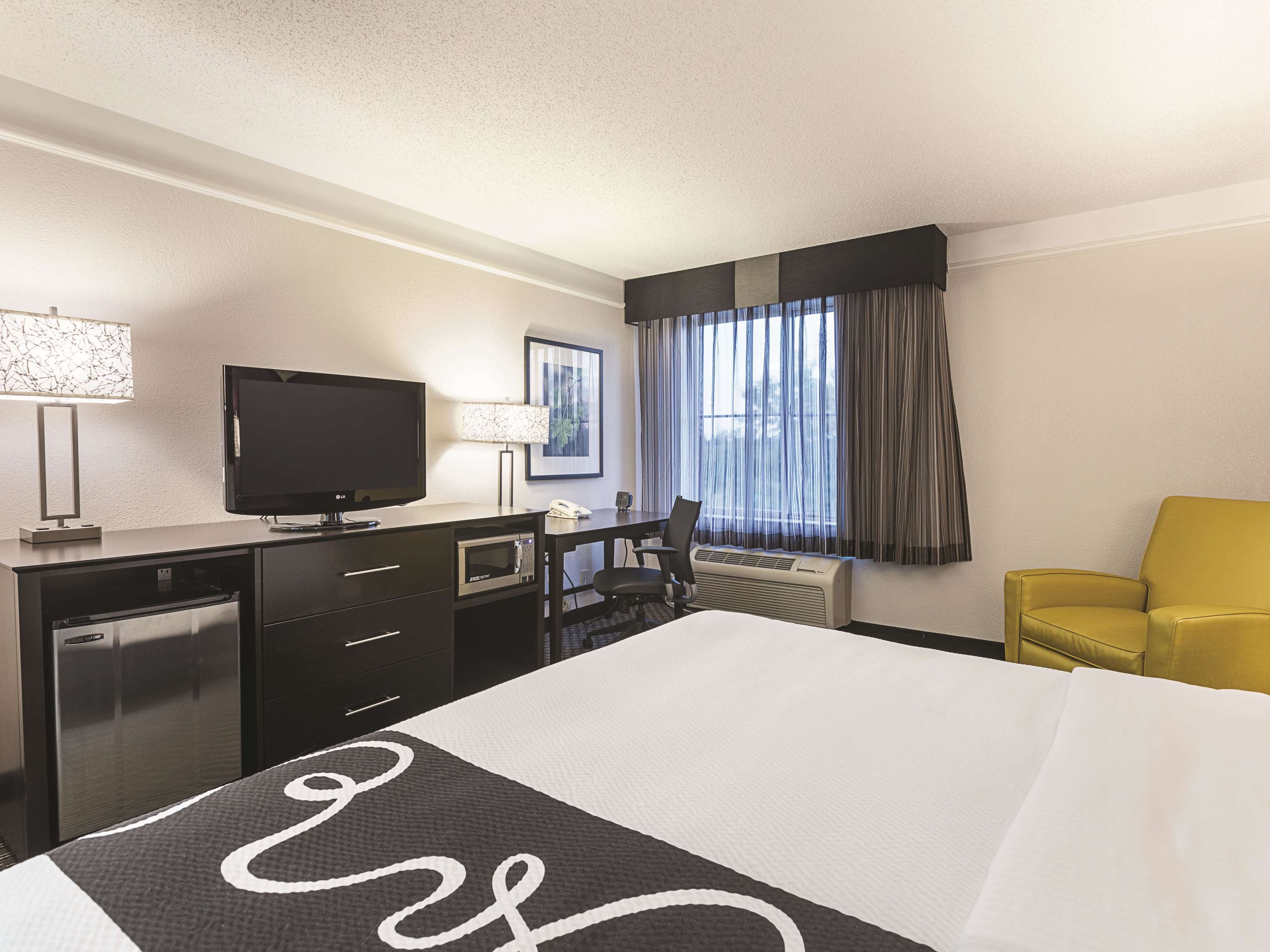 A hotel room with a bed, a desk with a television, a lamp, a chair, and a window with curtains.
