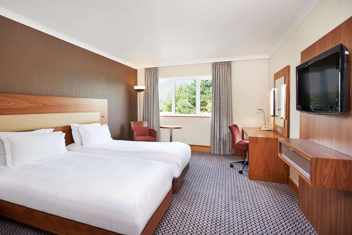 A hotel room with two beds, a desk, a chair, a TV, and a window with curtains.