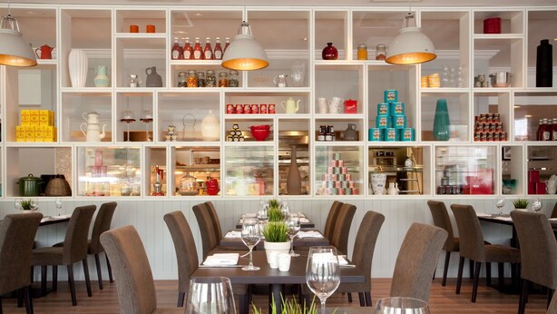 Uma sala de jantar com uma mesa comprida posta para seis pessoas, rodeada de cadeiras, e prateleiras exibindo diversos utensílios de cozinha e garrafas ao fundo.
