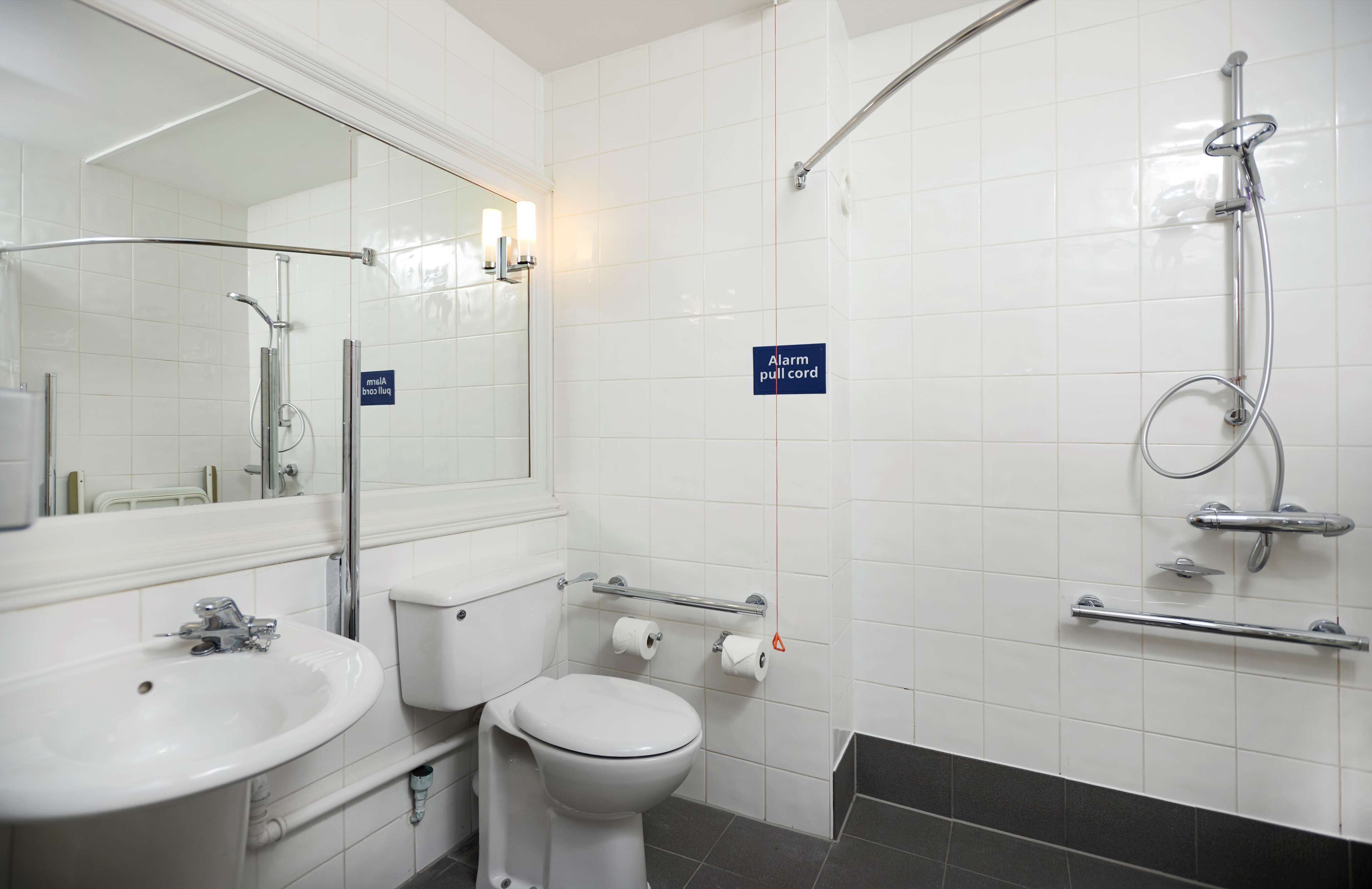 A bathroom with a sink, toilet, and shower. There is a sign indicating an alarm pull cord.