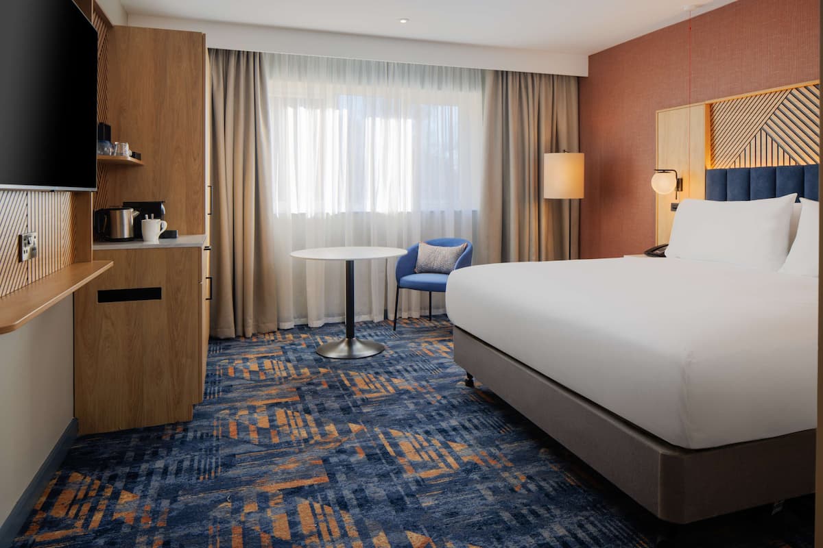 A hotel room with a large bed, a desk with a flat-screen TV, a small round table, a chair, and a window with curtains.