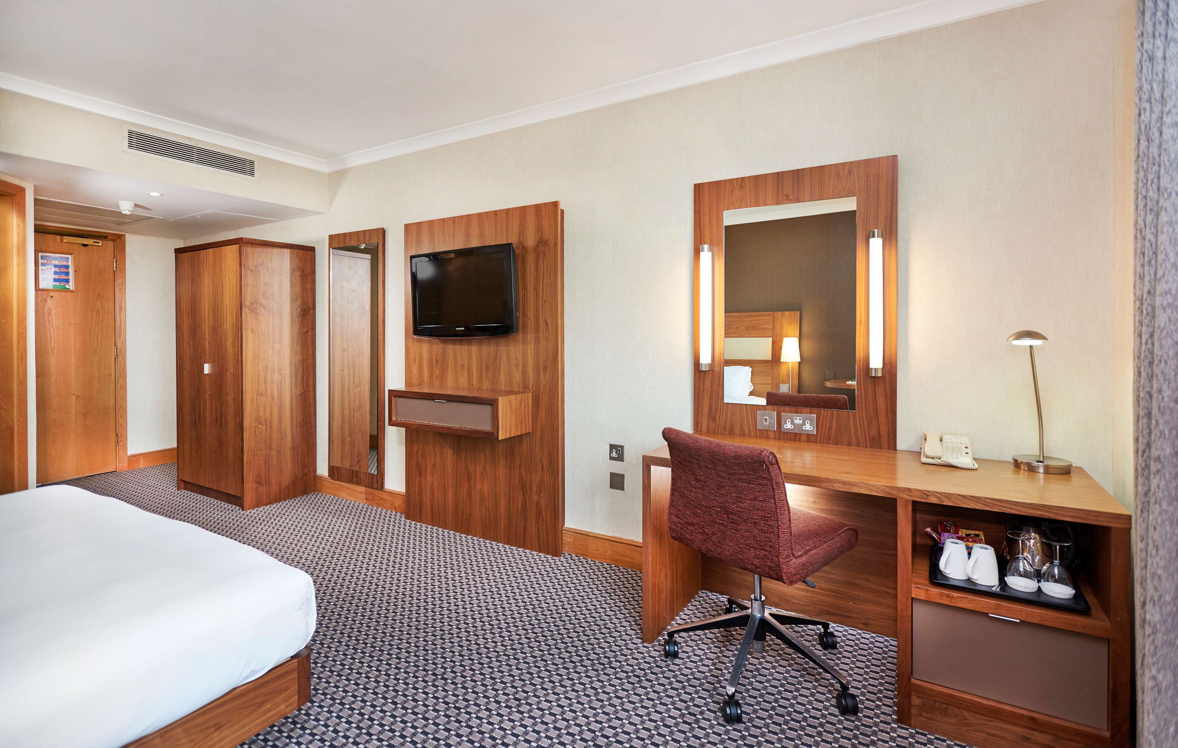 Une chambre d’hôtel comprenant un lit, une armoire en bois, un bureau avec un miroir, une télévision et une lampe de bureau.