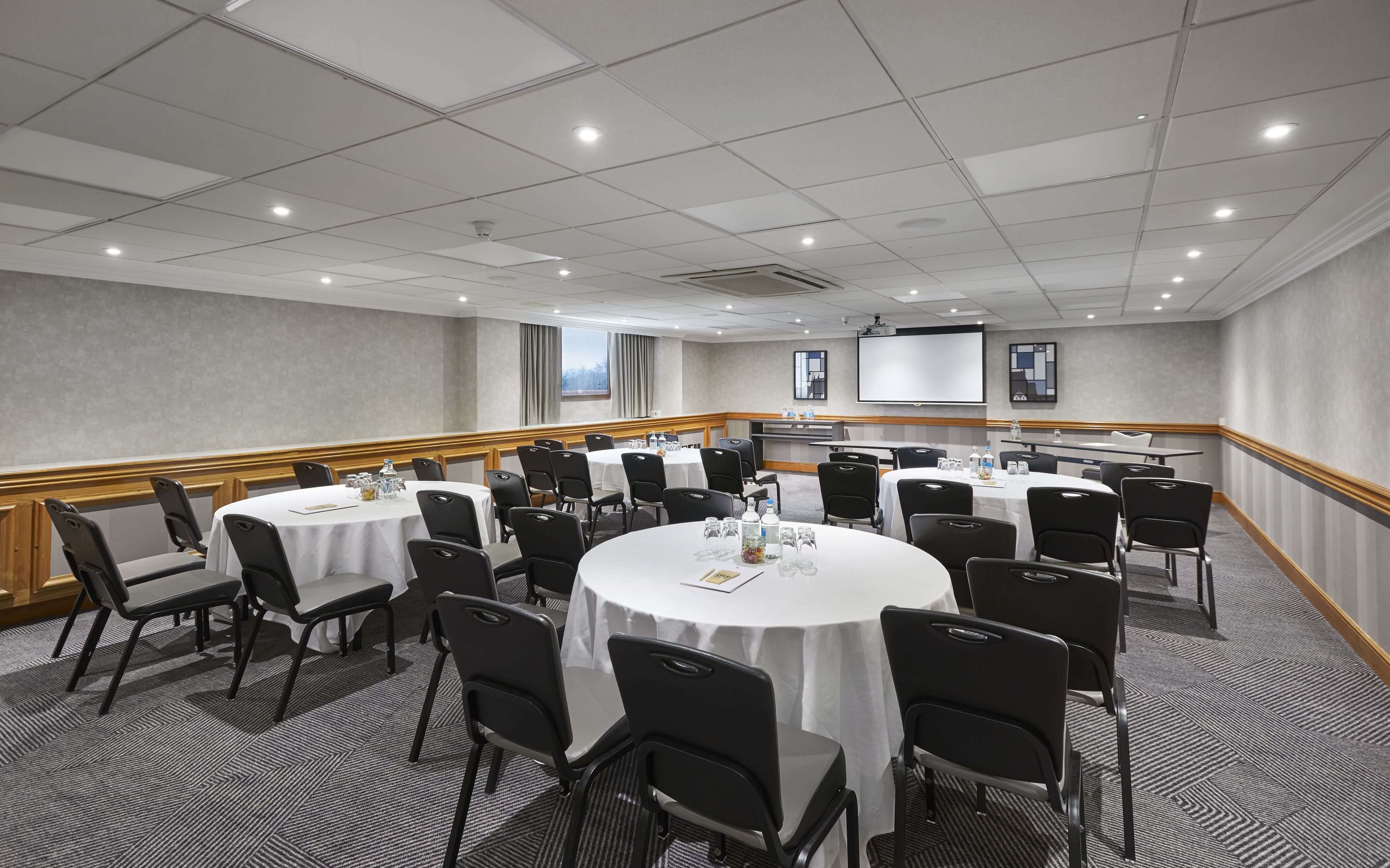 A conference room with round tables, black chairs, and a projector screen on the wall.