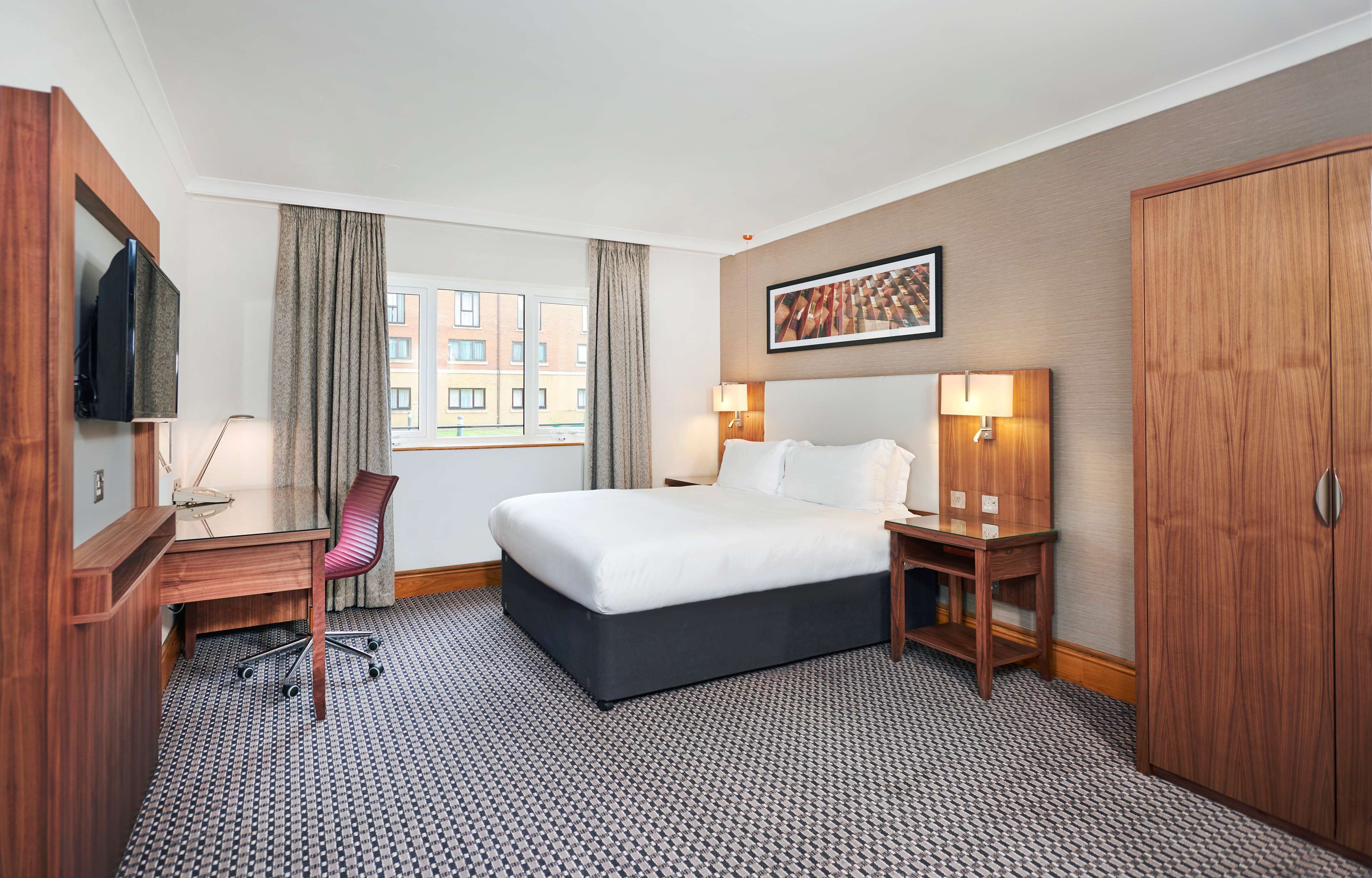 A hotel room with a bed, desk, chair, TV, and a large window with curtains.