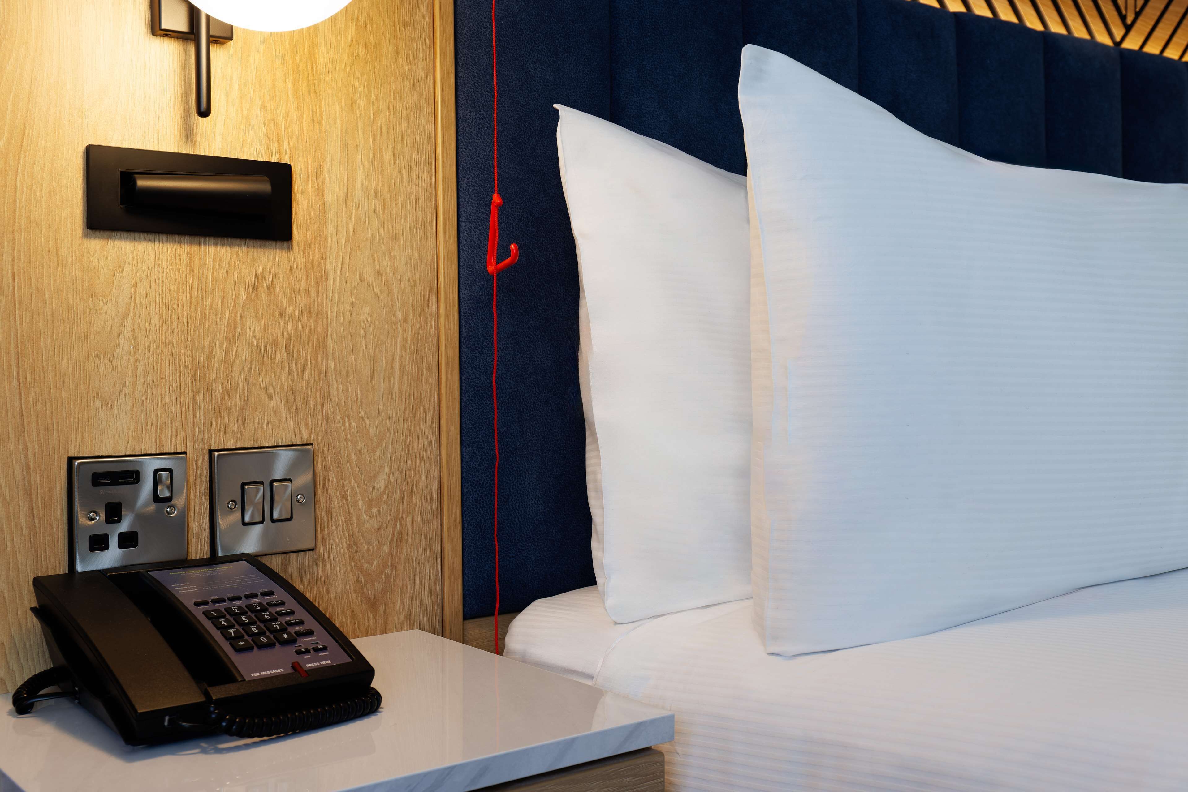 Una habitación de hotel con una cama, una mesita de noche con teléfono y una lámpara de pared.