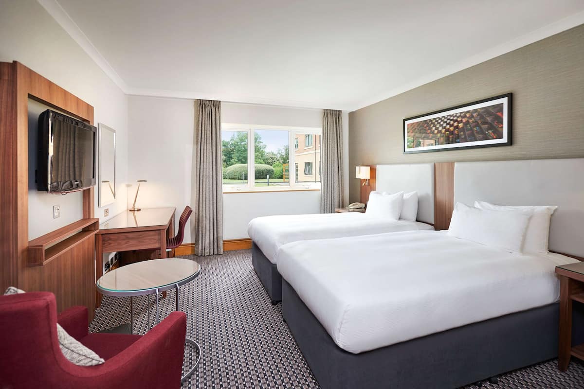A hotel room with two beds, a TV, a desk, a chair, and a window with a view of buildings.