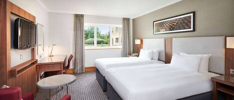 A hotel room with two beds, a TV, a desk, a chair, and a window with a view of buildings.