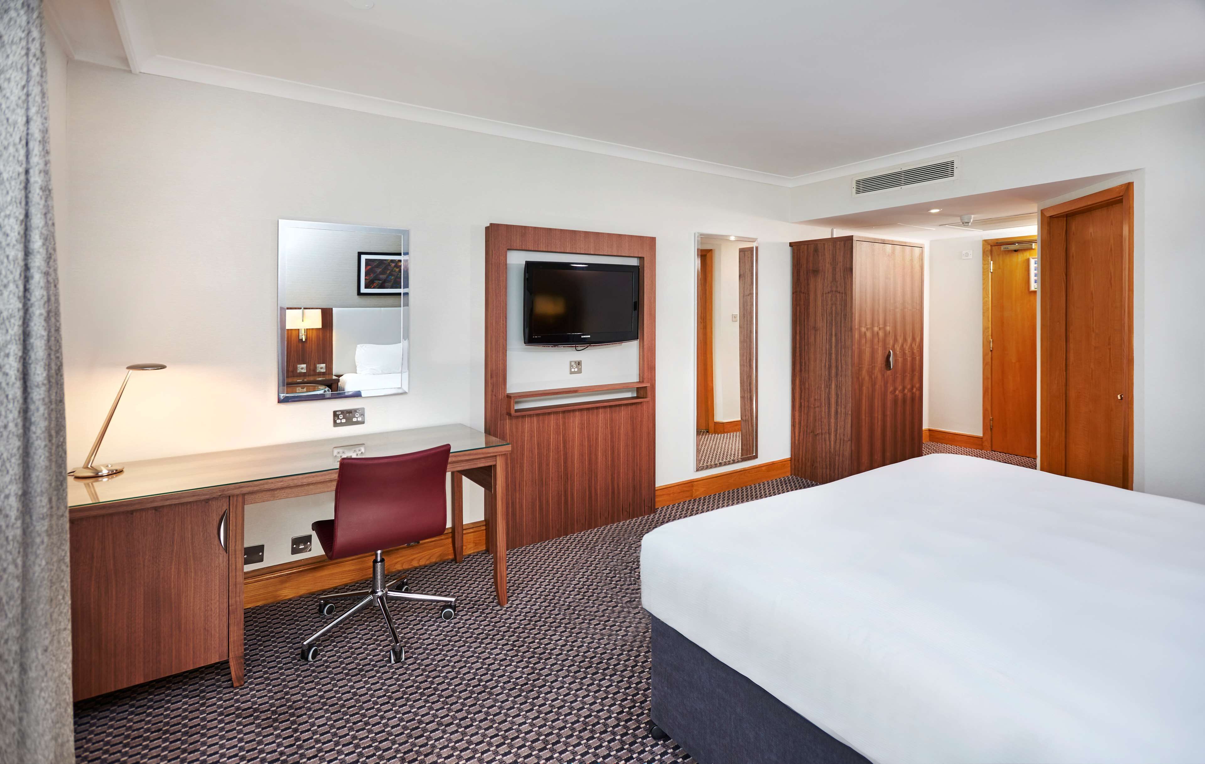 A hotel room with a bed, desk, chair, TV, wardrobe, and a mirror.