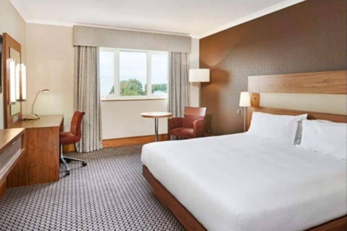 A hotel room with a large bed, a desk, a chair, a small table, and a window with curtains.