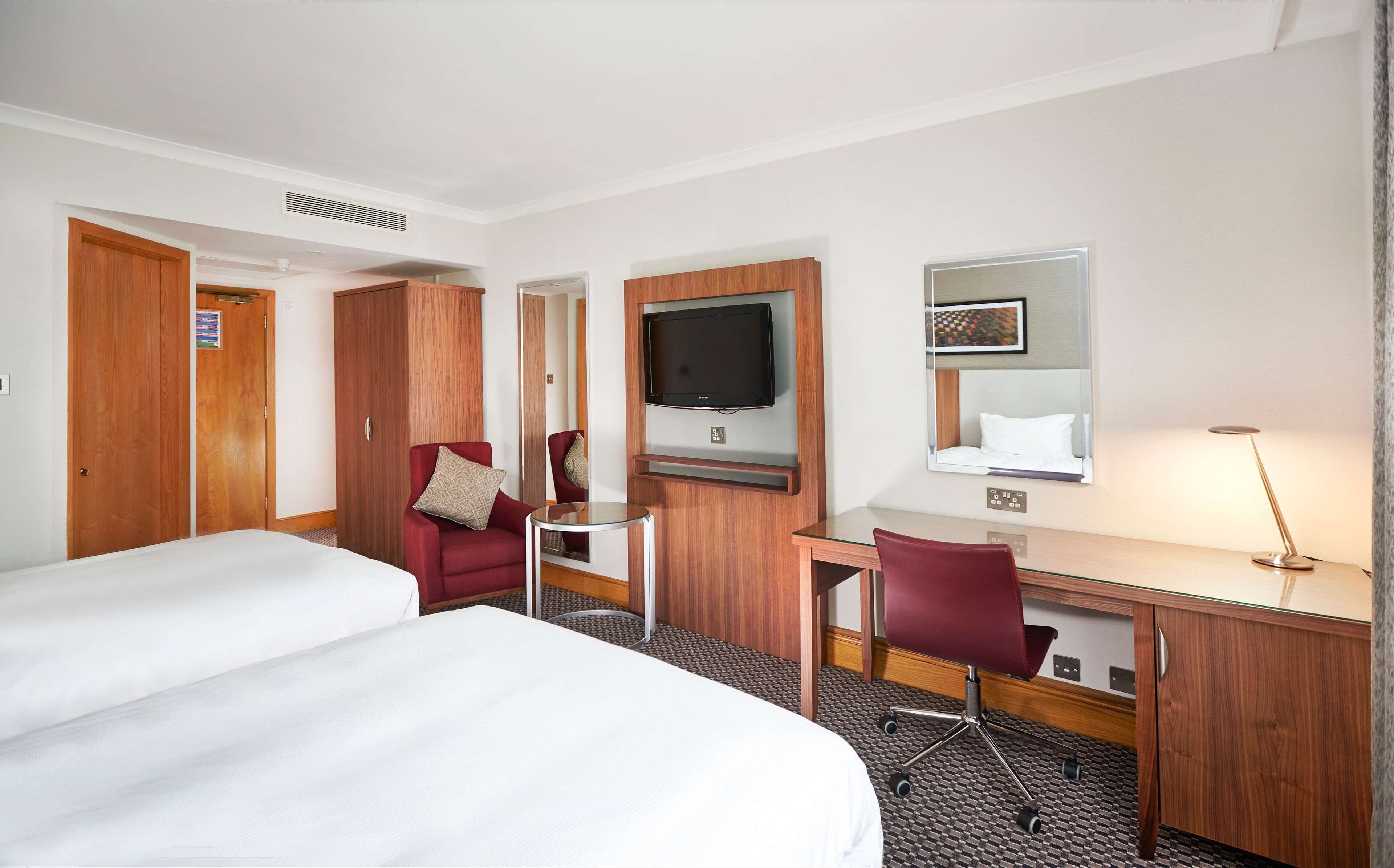 Una habitación de hotel con dos camas, un escritorio con silla, televisión, espejo y lámpara.