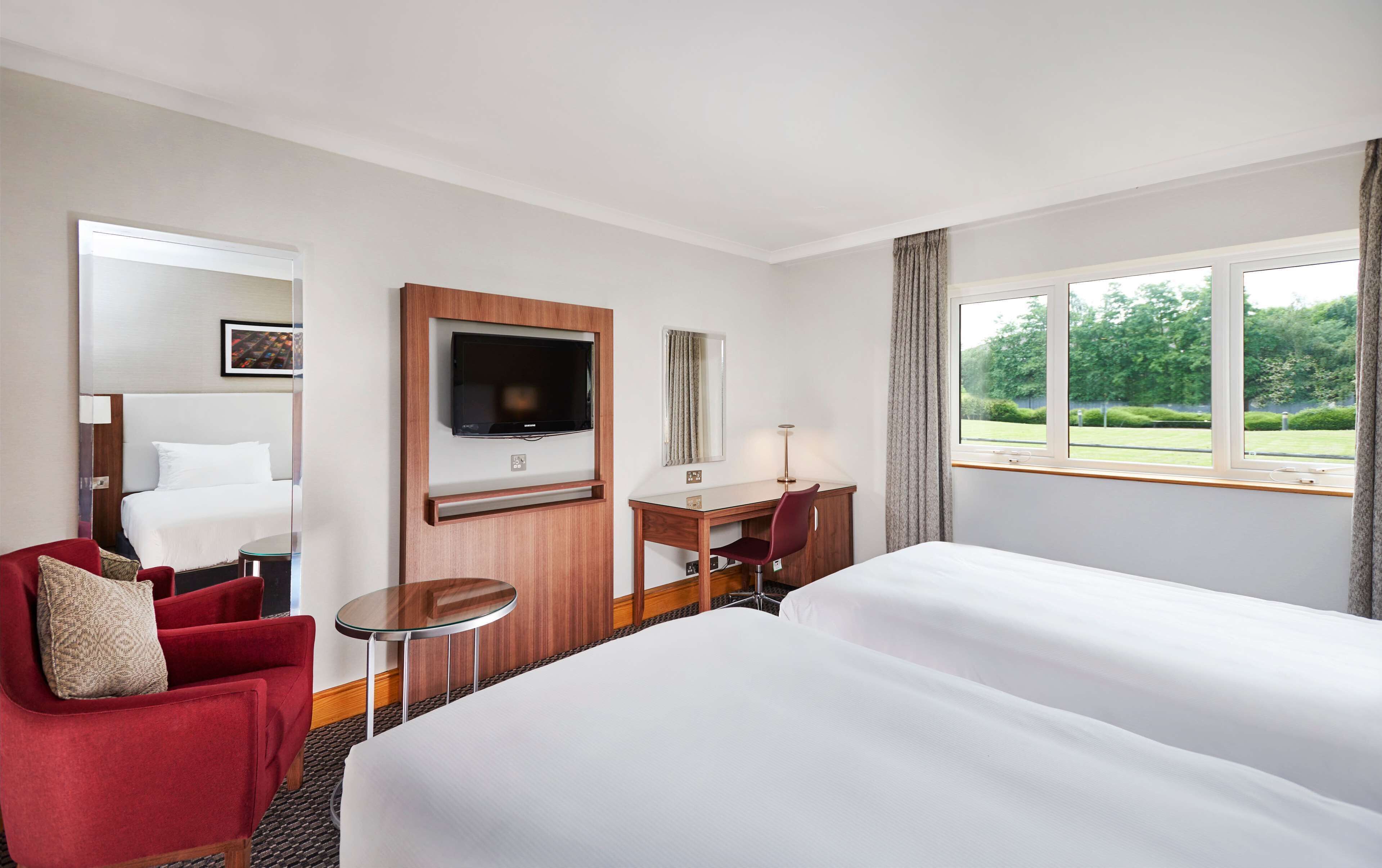 A hotel room with a large bed, a desk with a chair, a television, and a window with a view of greenery.