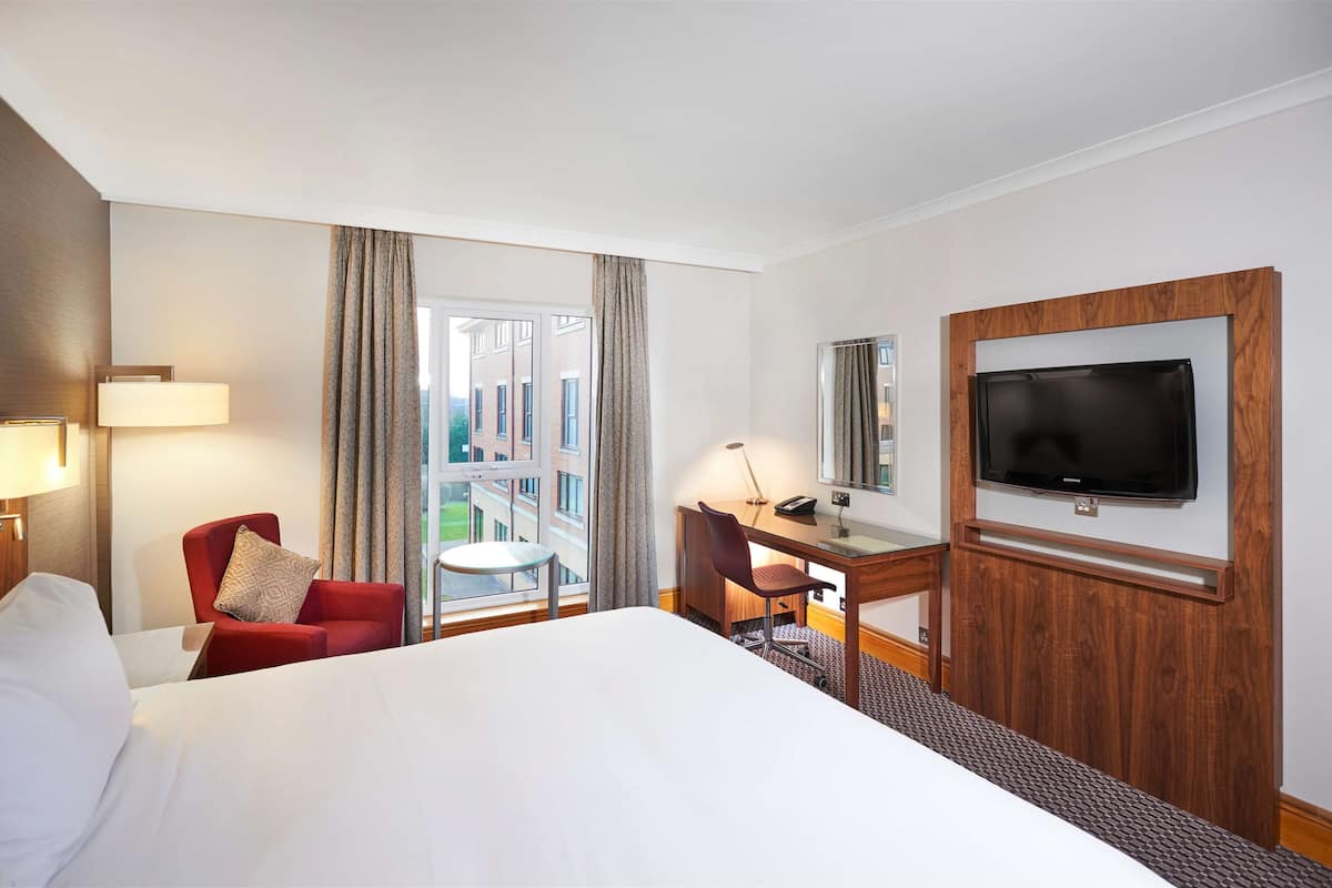 A hotel room with a large bed, a desk with a chair, a television mounted on the wall, and a window with curtains.
