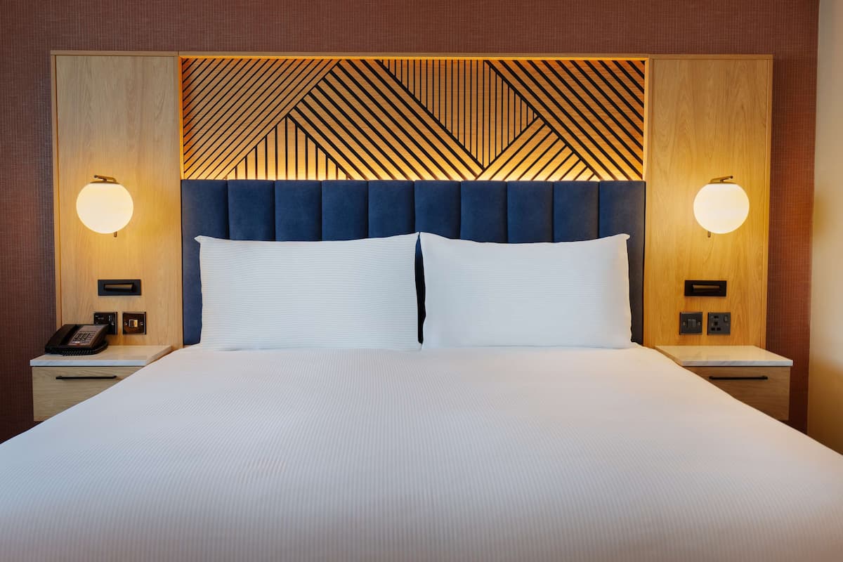 A hotel room with a large bed, two bedside tables, a wooden headboard with a geometric pattern, and wall-mounted lamps.