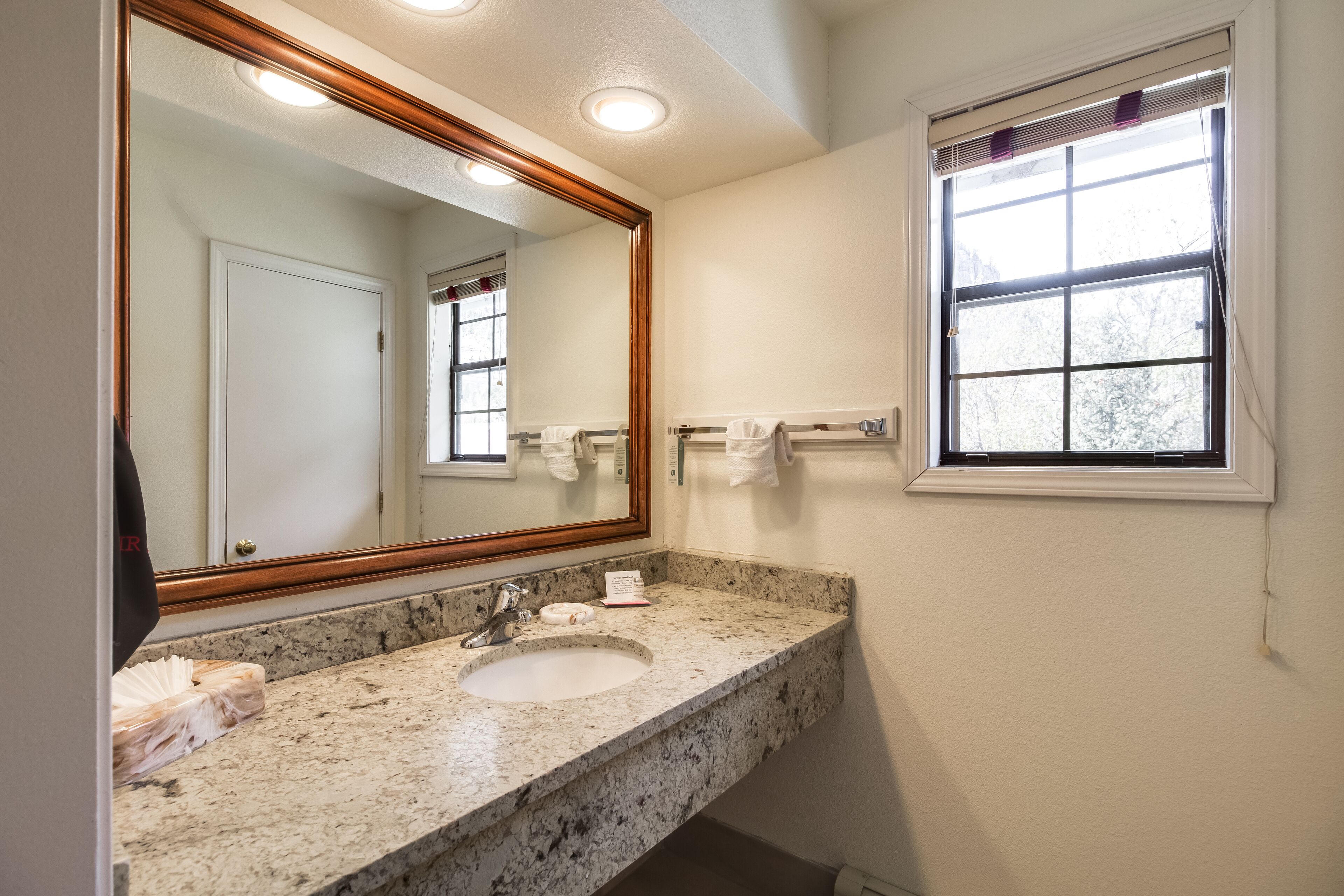 Une salle de bain avec un lavabo doté d’un plan de travail en granit, un grand miroir et une fenêtre avec des stores.
